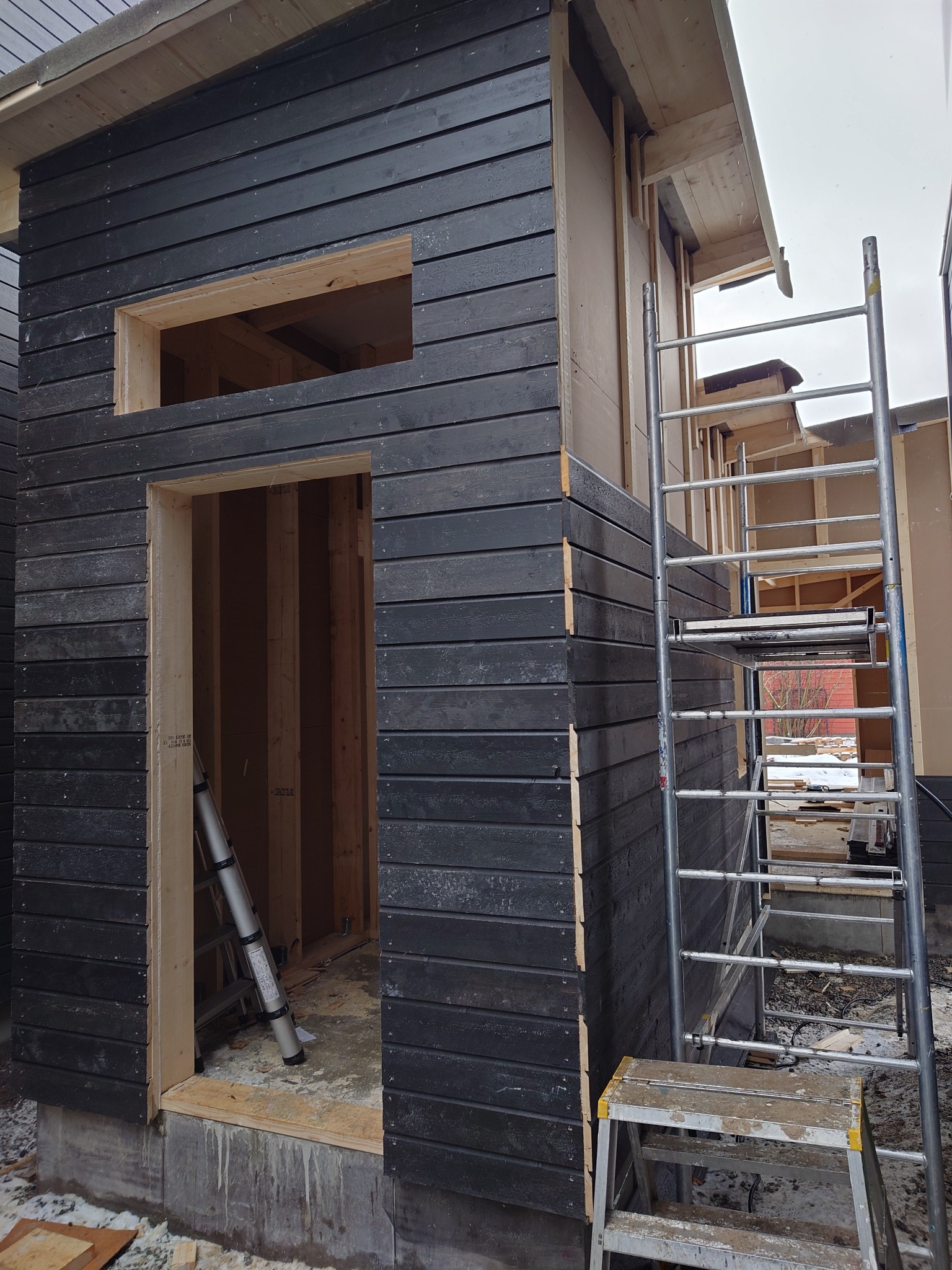 Surowa elewacja z ciemnego drewna w trakcie budowy, widoczne ramy okienne i drzwiowe, rusztowanie aluminiowe i drabina, teren budowy z resztkami materiałów.