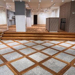 TCC TOMASZ CHARKOT COMPANY - Wnętrze sklepu po remoncie: dekoracyjna podłoga z drewnianymi obramowaniami i szarym kamieniem, drewniane schody, ściany z pionowych paneli, oświetlenie punktowe na suficie, szare filary z białymi...