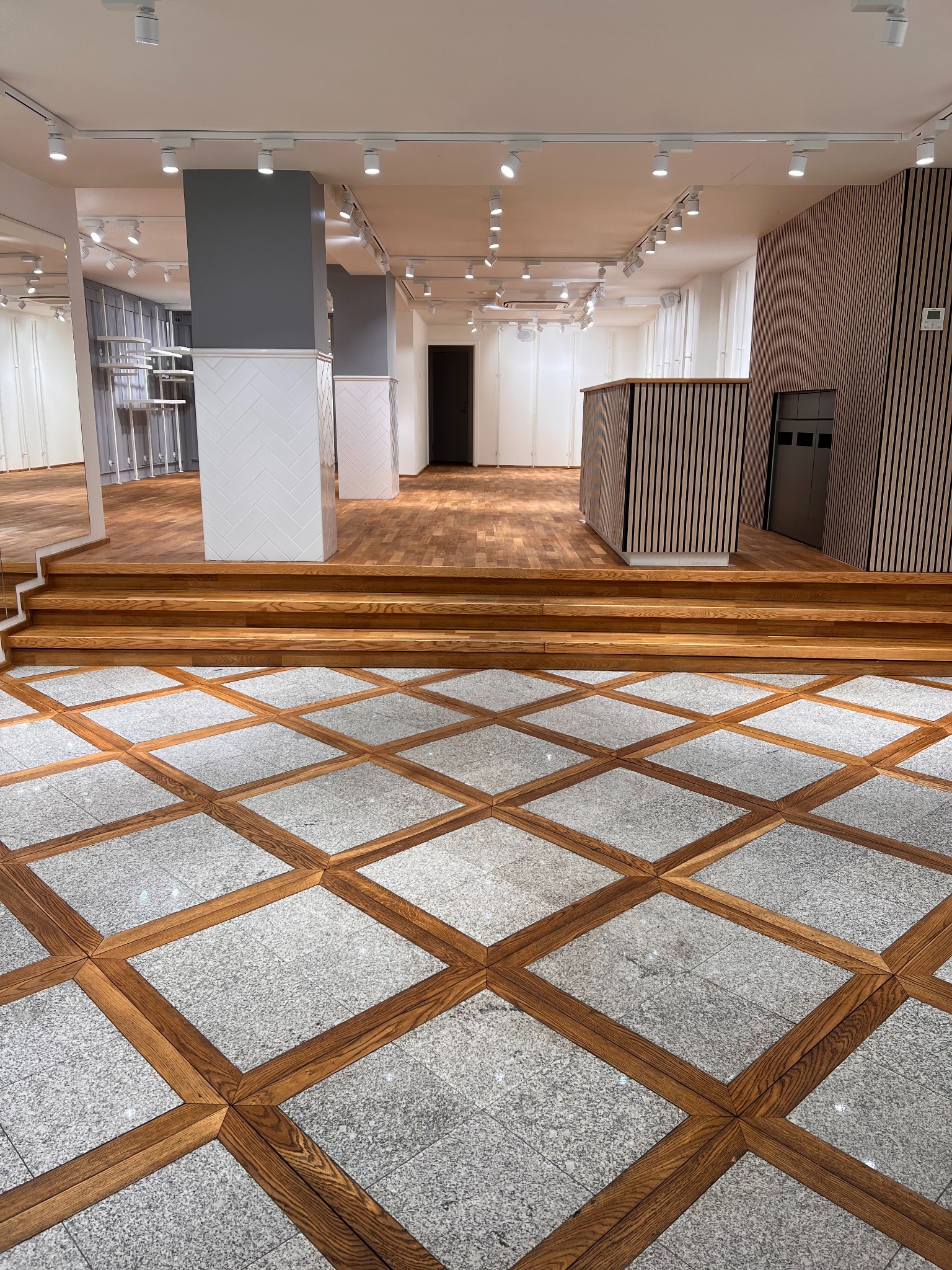 Wnętrze sklepu po remoncie: dekoracyjna podłoga z drewnianymi obramowaniami i szarym kamieniem, drewniane schody, ściany z pionowych paneli, oświetlenie punktowe na suficie, szare filary z białymi...