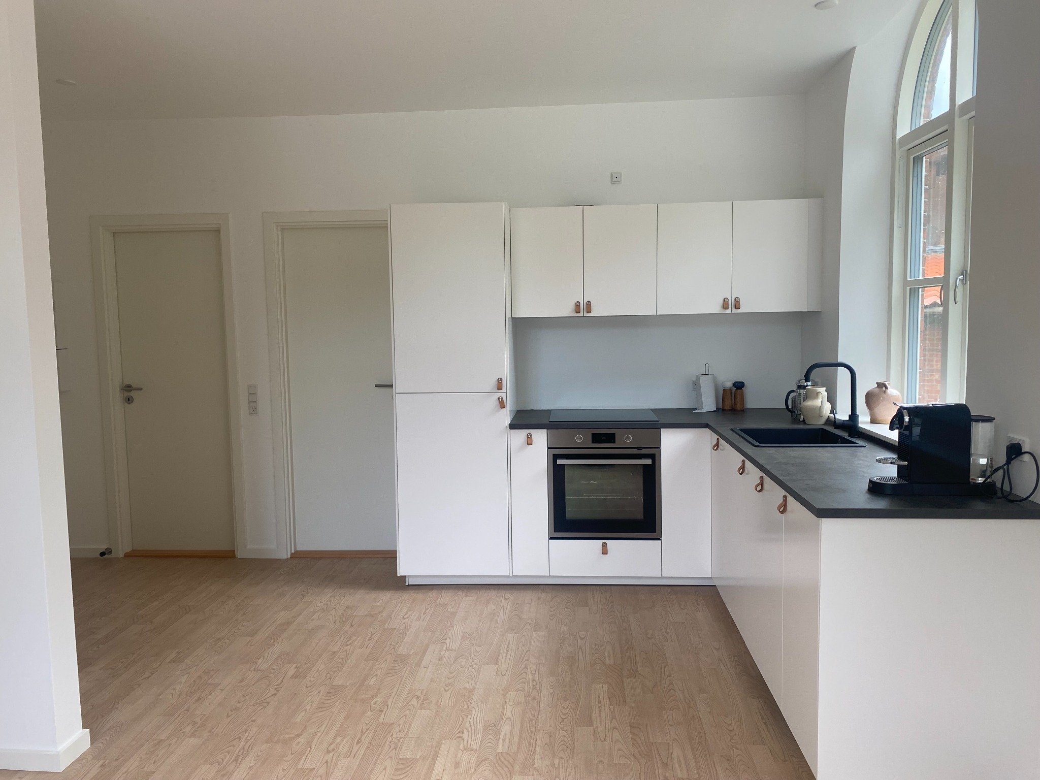 Jasna kuchnia z białymi szafkami, czarnym blatem i drewnianą podłogą; wysokie okno łukowe w tle.