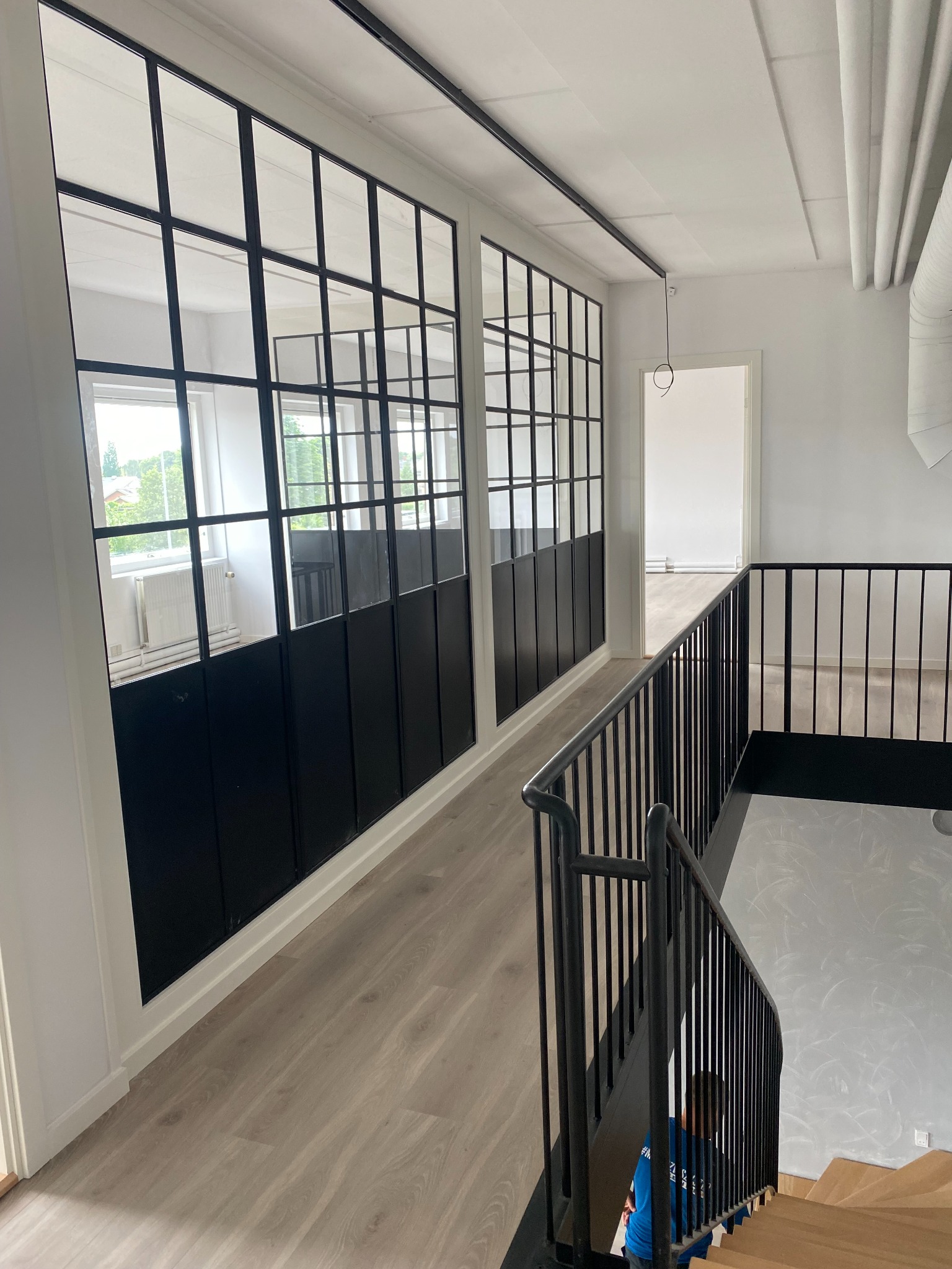 Nowoczesne wnętrze z przeszkloną ścianą działową w czarnej ramie, drewnianą podłogą i schodami z czarną balustradą, widok z góry na osobę schodzącą po schodach.