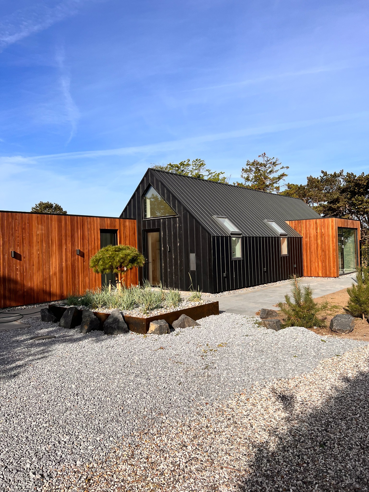 Nowoczesny dom jednorodzinny z czarną elewacją i drewnianymi panelami, otoczony żwirowym podłożem i minimalistyczną roślinnością.