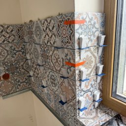 Tomasz Wojciechowski - Układanie dekoracyjnych płytek ściennych z użyciem systemu poziomowania. Widoczne plastikowe klipsy i kliny. Fragment okna z drewnianą ramą w tle.