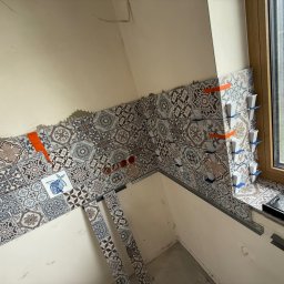 Tomasz Wojciechowski - Układanie dekoracyjnych płytek ściennych w łazience, z użyciem poziomowania. Widoczne narzędzia glazurnicze i fragment okna. Płytki z motywem patchwork.
