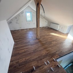 Tomasz Wojciechowski - Podłoga na poddaszu z brązowych płytek imitujących drewno, z białymi krzyżykami dystansowymi. Widoczny fragment okna i drewniany słup konstrukcyjny.