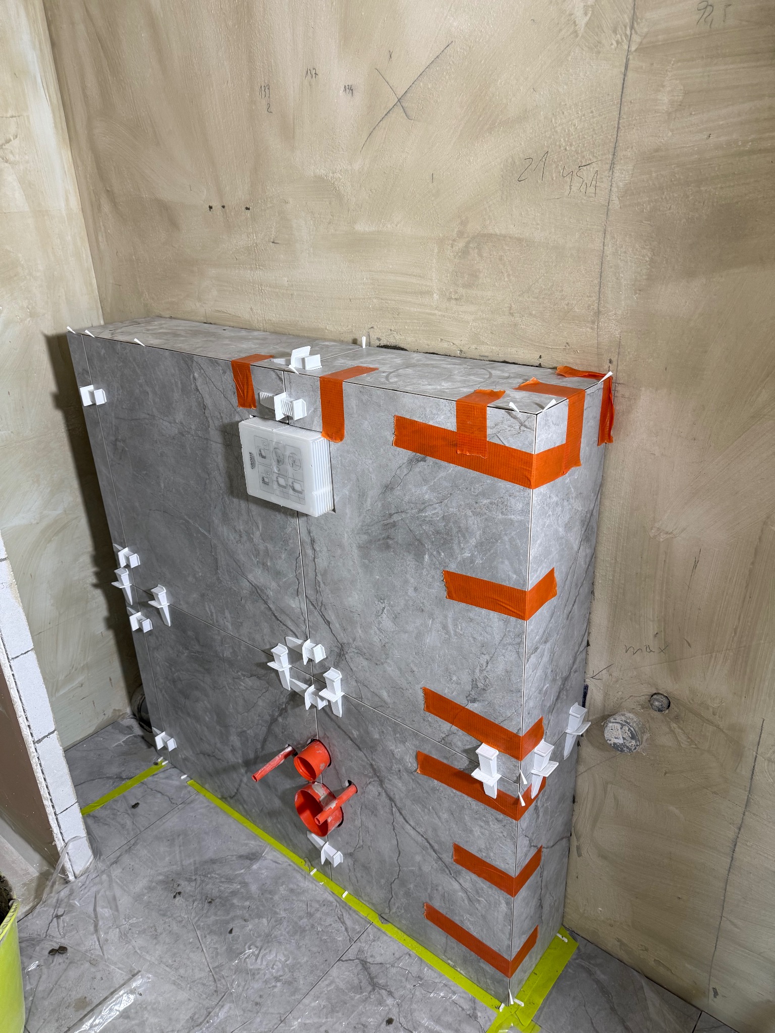 Obudowa stelaża WC z szarych płytek imitujących marmur, zabezpieczona pomarańczową taśmą i białymi krzyżykami dystansowymi. Widoczne fragmenty łazienki w trakcie prac.