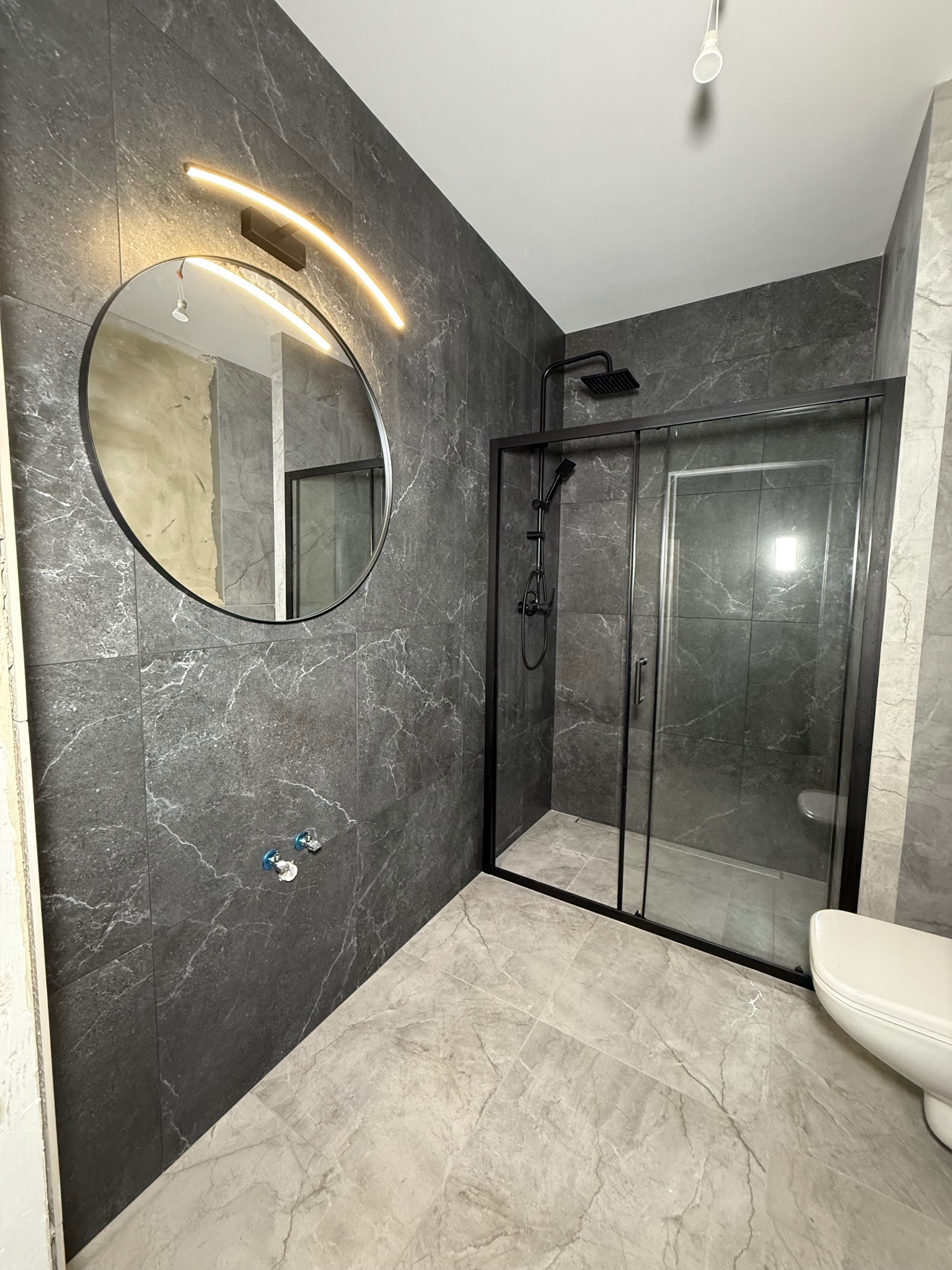 Nowoczesna łazienka w szarościach z kabiną prysznicową typu walk-in, okrągłym lustrem z podświetleniem LED i marmurowymi płytkami na ścianach i podłodze.