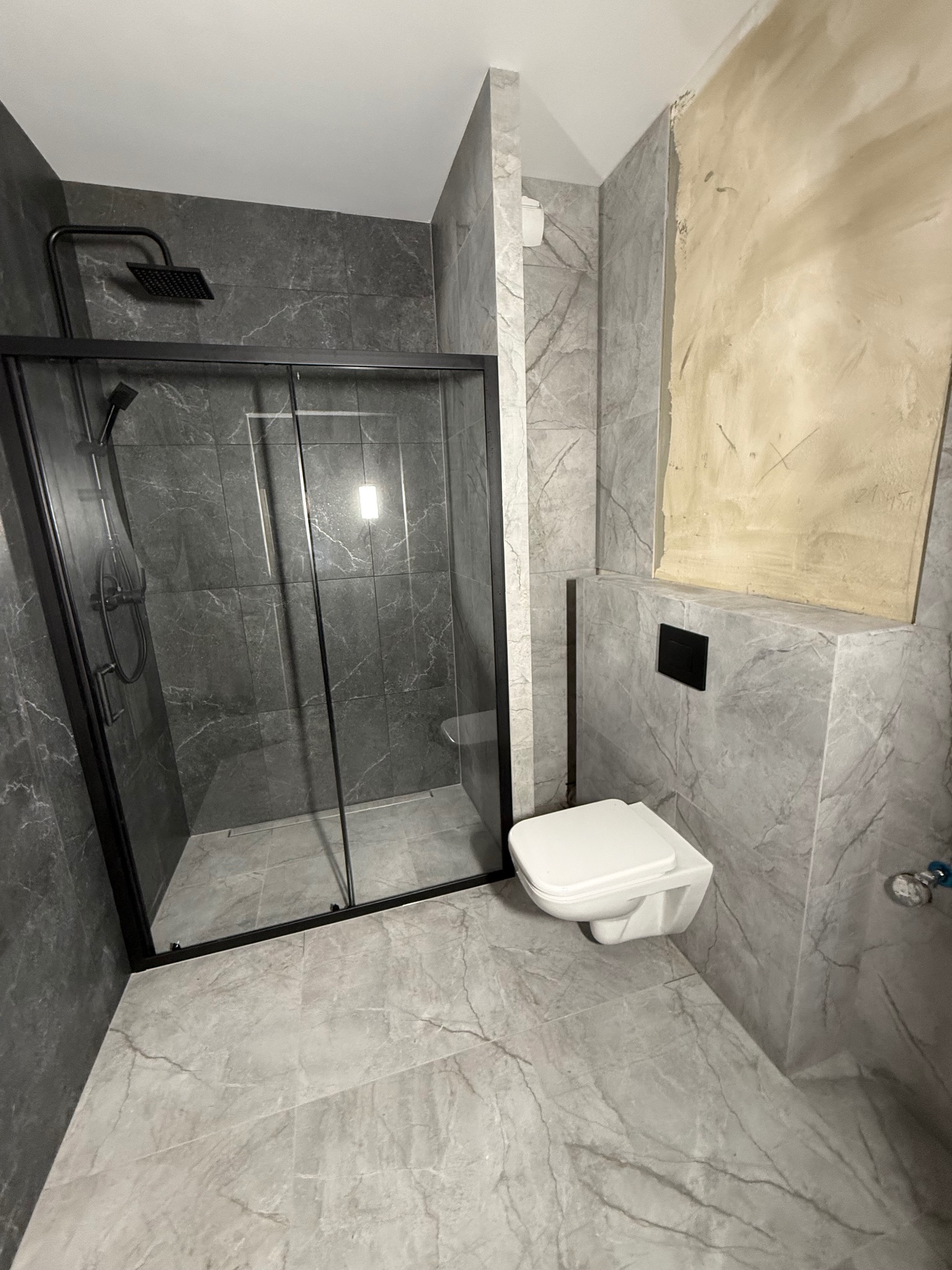 Nowoczesna łazienka w szarościach z kabiną prysznicową typu walk-in z czarnymi profilami i podwieszaną toaletą. Częściowo wykończona ściana z widocznym surowym tynkiem.