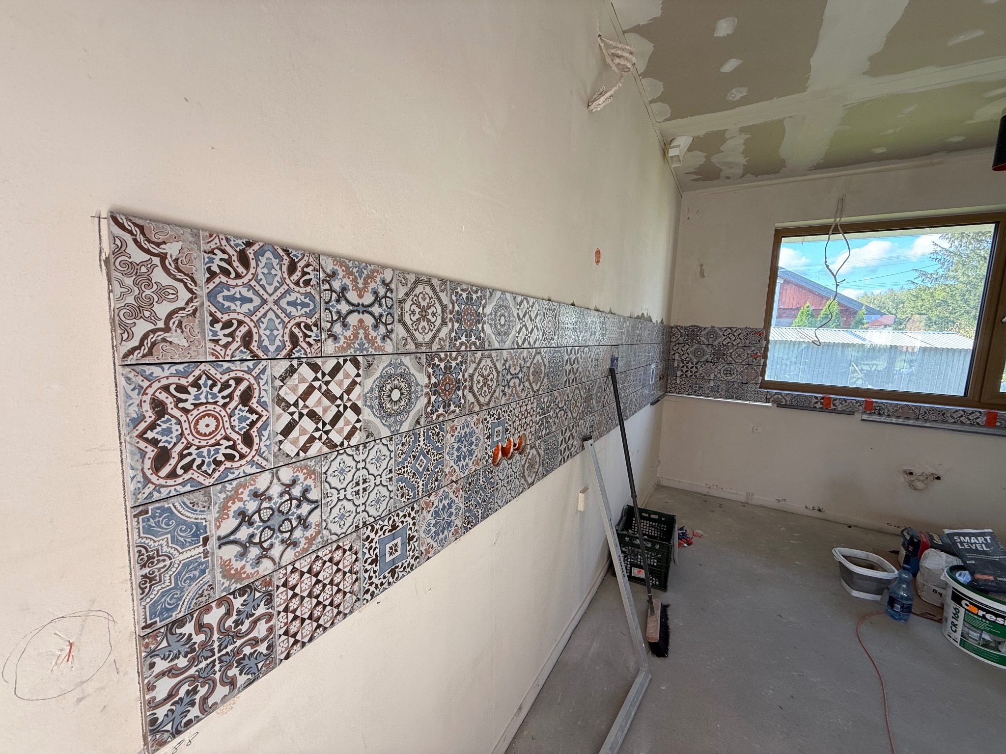 Fragment ściany z ułożonymi, wzorzystymi płytkami w trakcie prac wykończeniowych. Widoczne narzędzia i materiały budowlane. Częściowo wykończone wnętrze.