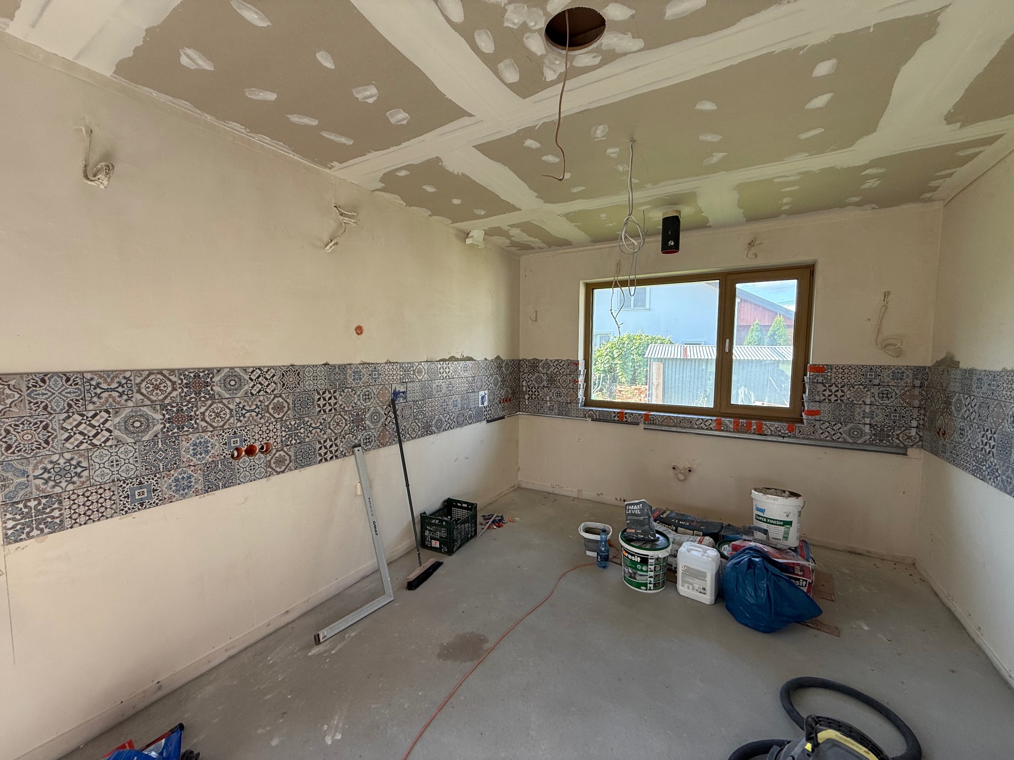 Wnętrze w trakcie prac: częściowo ułożone płytki dekoracyjne, szpachlowana sufit, widoczne przewody elektryczne, narzędzia i materiały budowlane. Ujęcie z poziomu podłogi.