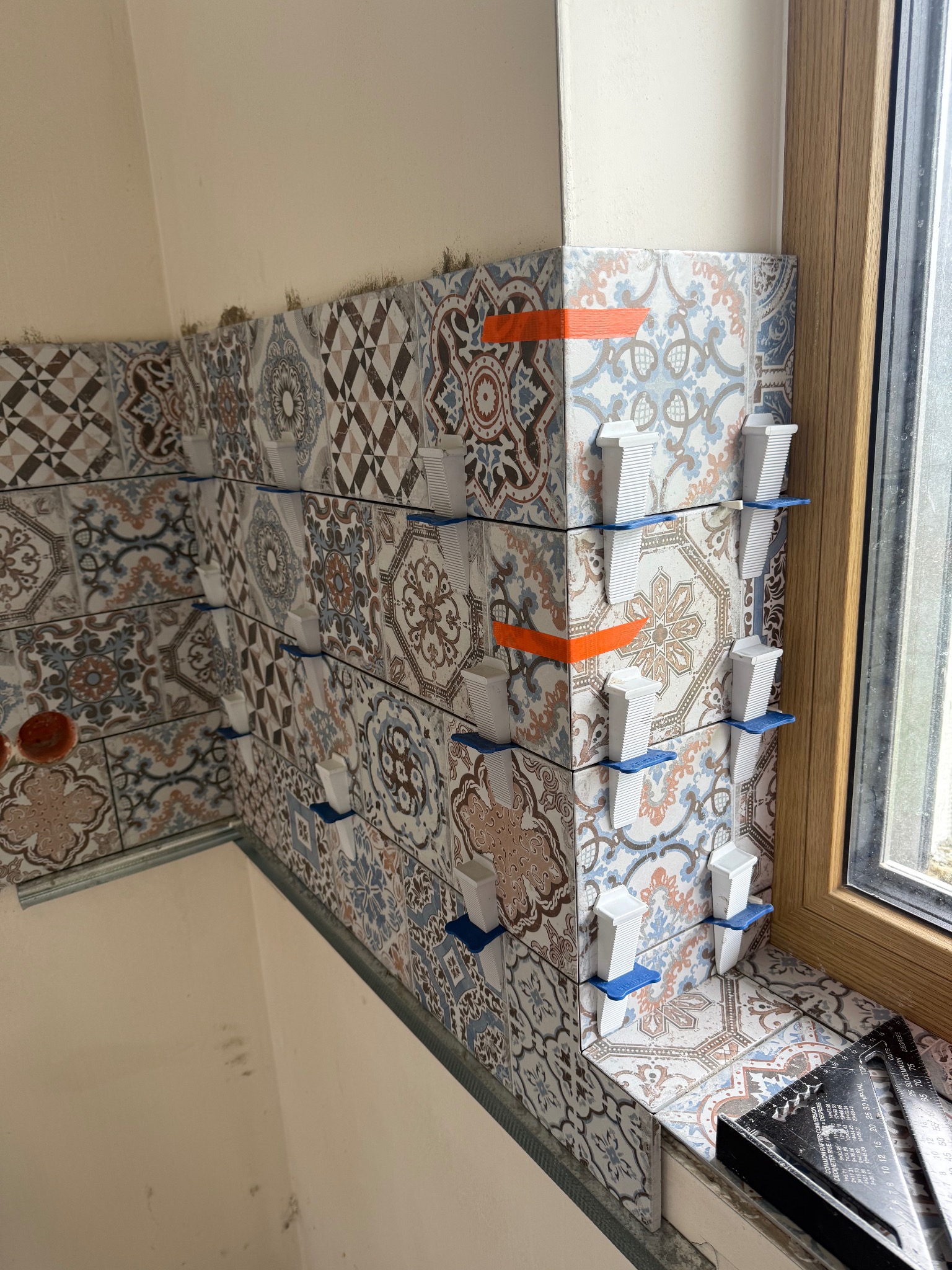 Układanie dekoracyjnych płytek ściennych z użyciem systemu poziomowania. Widoczne plastikowe klipsy i kliny. Fragment okna z drewnianą ramą w tle.