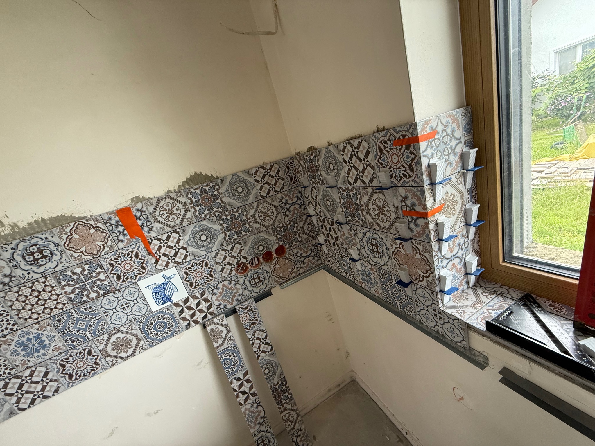 Układanie dekoracyjnych płytek ściennych w łazience, z użyciem poziomowania. Widoczne narzędzia glazurnicze i fragment okna. Płytki z motywem patchwork.