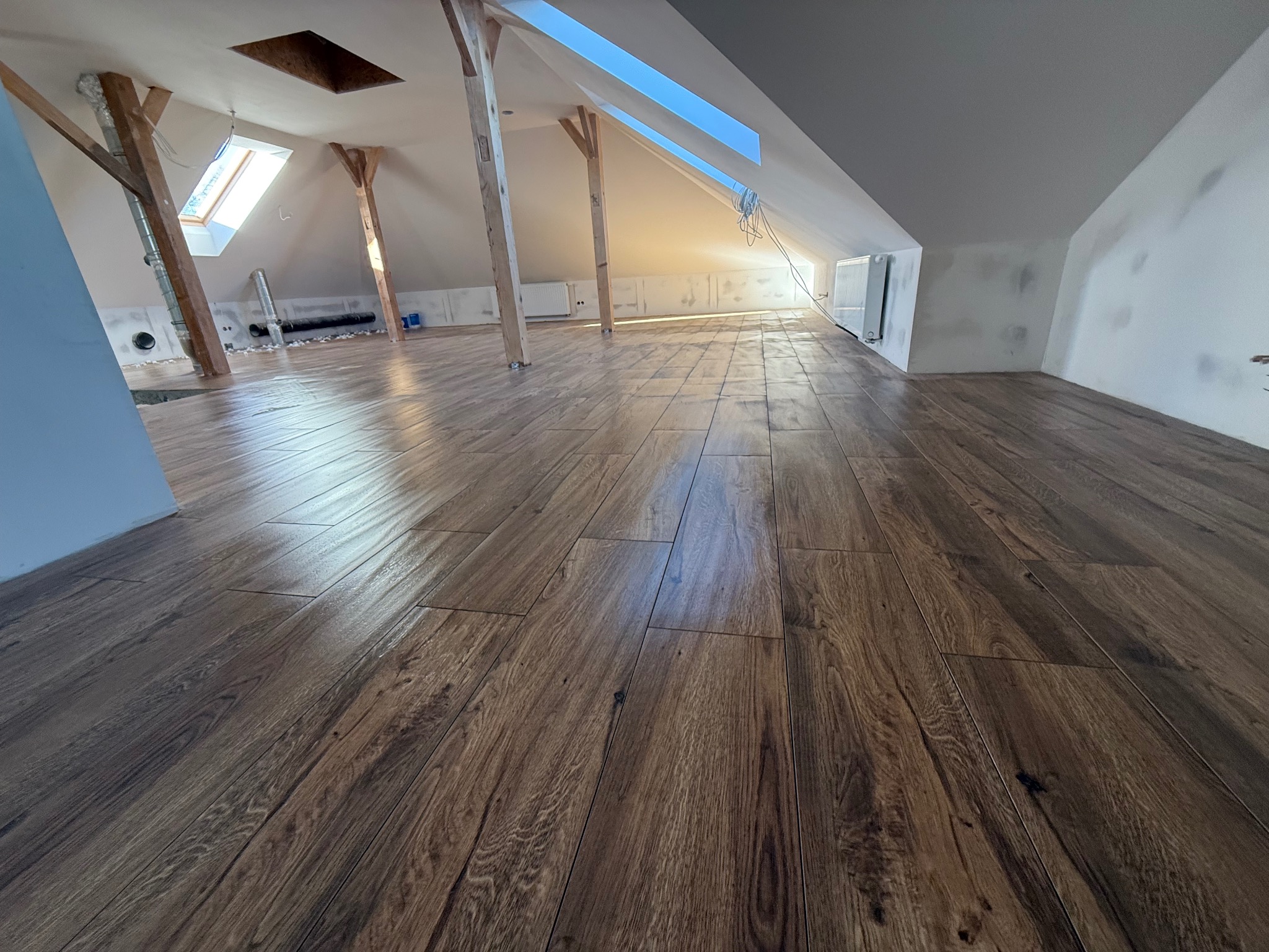 Ukończona podłoga z paneli imitujących drewno na poddaszu z oknami dachowymi i drewnianymi belkami konstrukcyjnymi. Widoczne grzejniki i ślady po szpachlowaniu ścian.