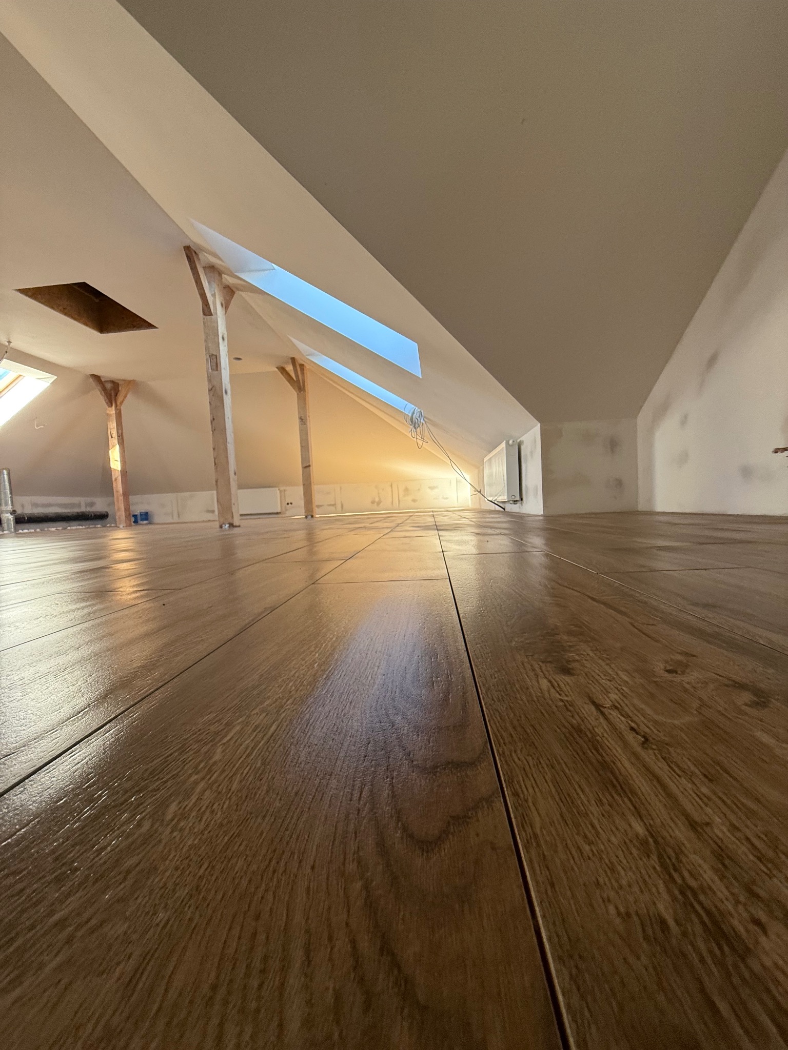 Ukończona podłoga z paneli drewnopodobnych na poddaszu z oknami dachowymi. Widok z niskiej perspektywy podkreśla wzór i połysk paneli. Ściany przygotowane do malowania.