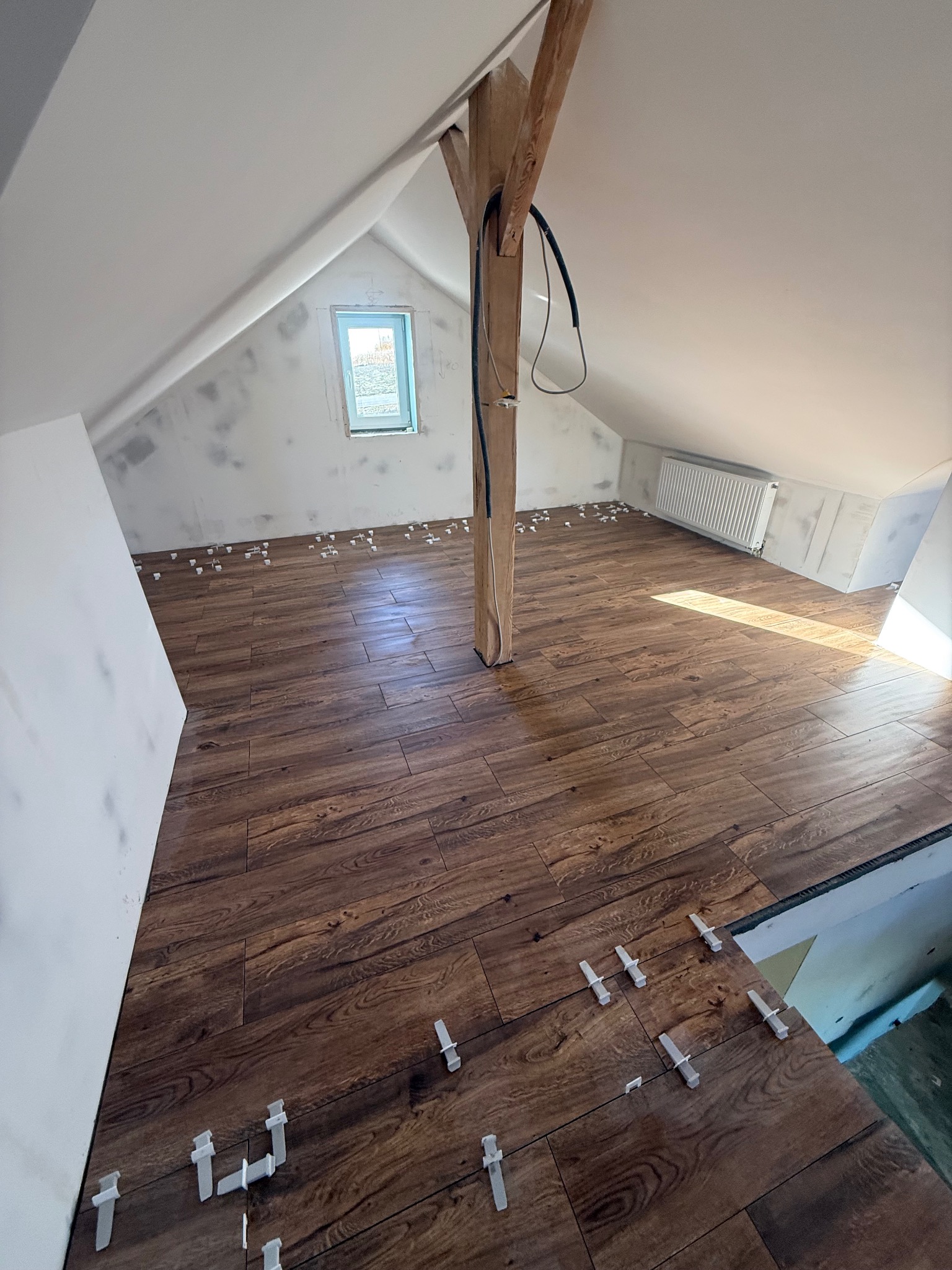 Podłoga na poddaszu z brązowych płytek imitujących drewno, z białymi krzyżykami dystansowymi. Widoczny fragment okna i drewniany słup konstrukcyjny.