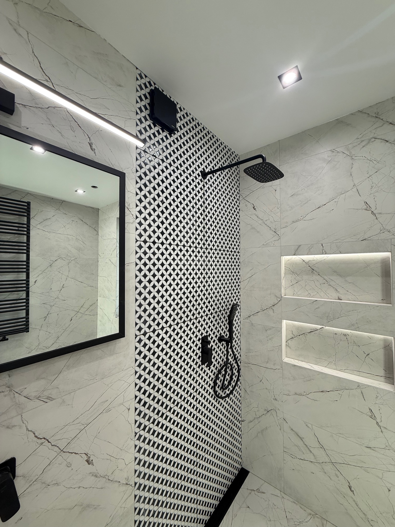 Nowoczesna łazienka z czarno-białą mozaiką, marmurowymi płytkami i czarną armaturą. Widoczne wnęki z podświetleniem i duże lustro.