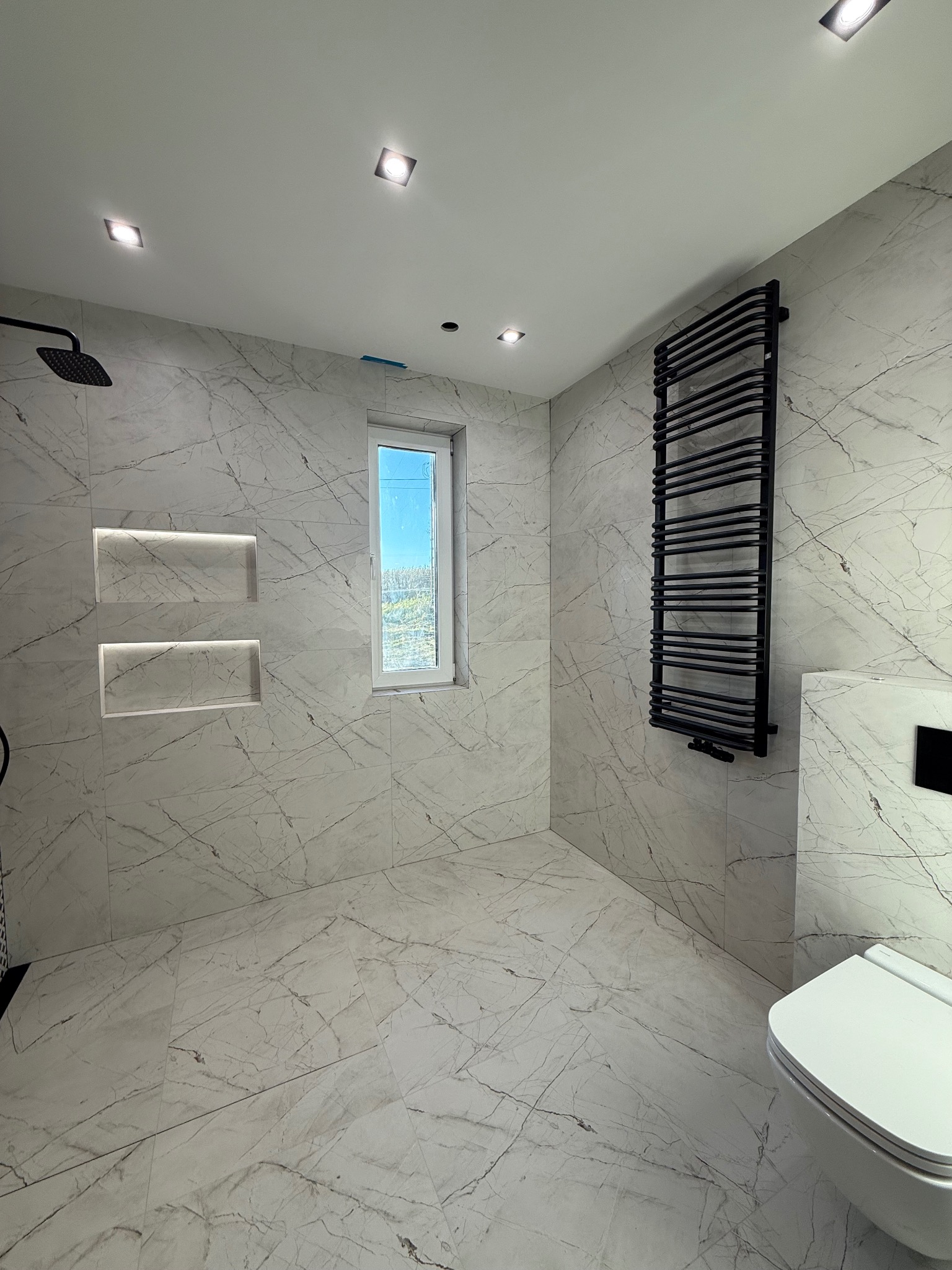 Elegancka łazienka z marmurowymi płytkami, czarnym grzejnikiem drabinkowym i wnękami z podświetleniem LED. Nowoczesny design z oknem i białym sanitariatem.