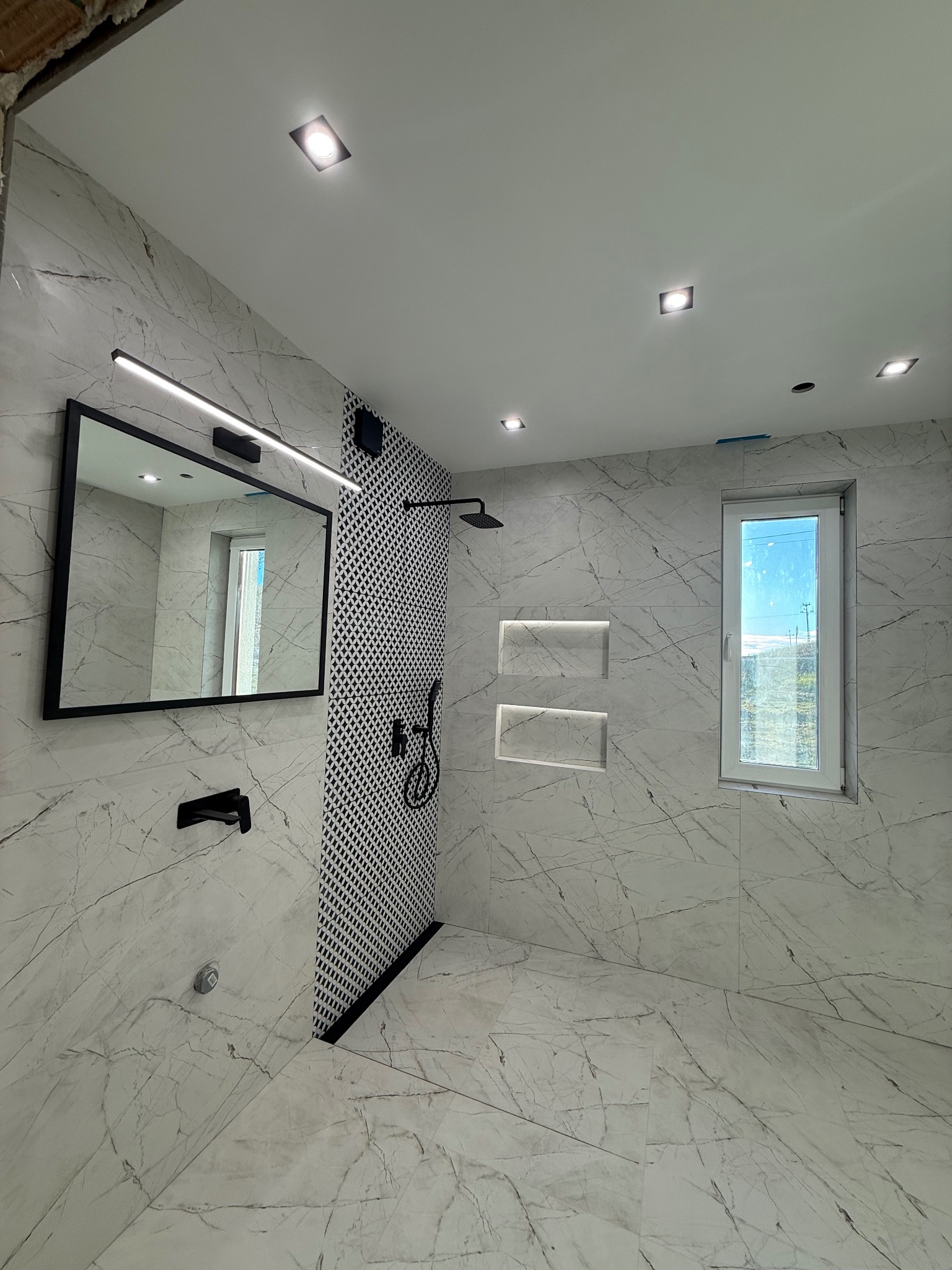 Nowoczesna łazienka z marmurowymi płytkami, czarną armaturą i geometrycznym wzorem na ścianie prysznicowej. Widoczne wnęki w ścianie i lustro z oświetleniem LED.