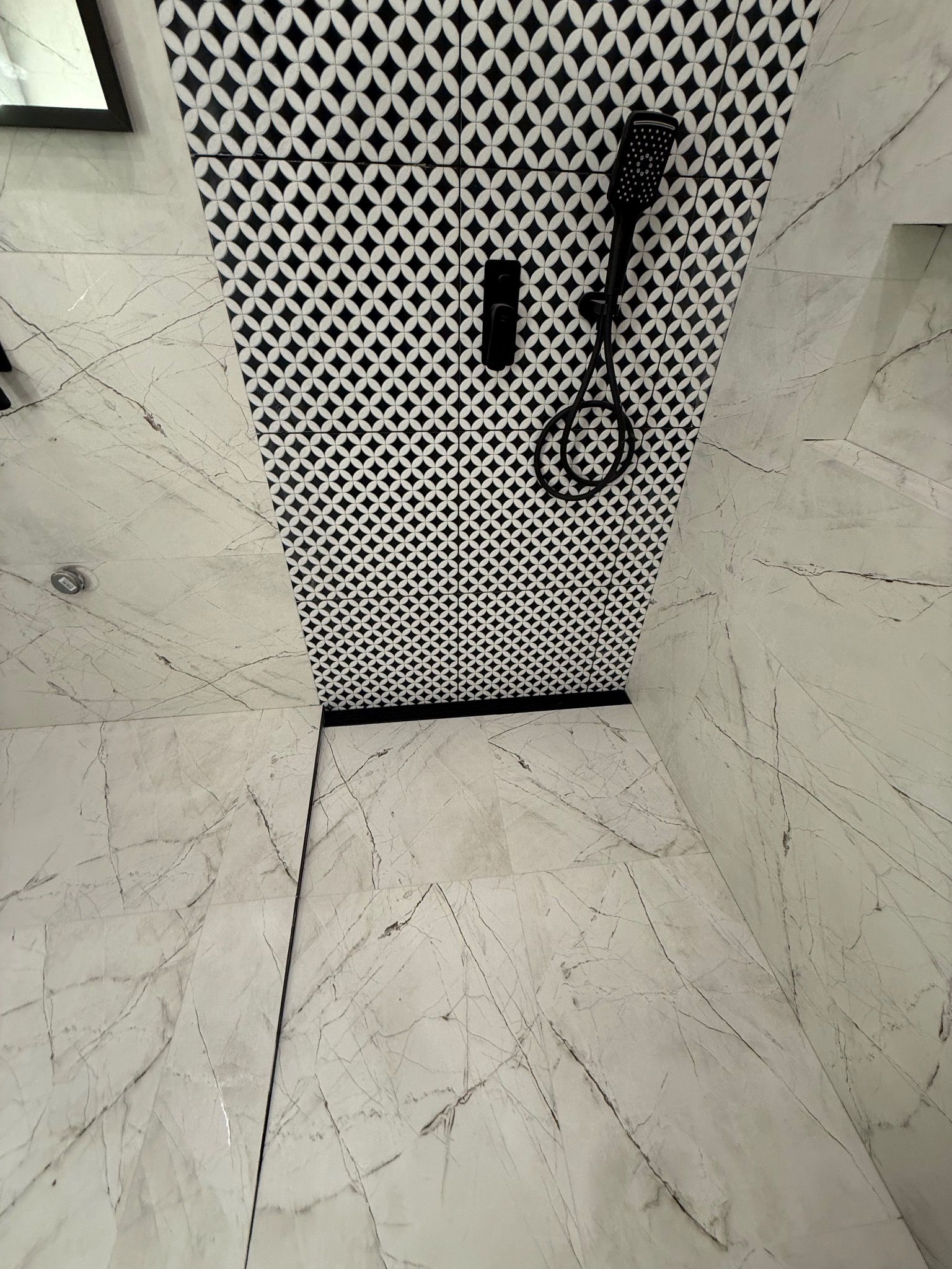 Nowoczesna łazienka z czarno-białymi płytkami o geometrycznym wzorze na ścianie prysznicowej i marmurowymi płytkami na podłodze. Czarny zestaw prysznicowy.