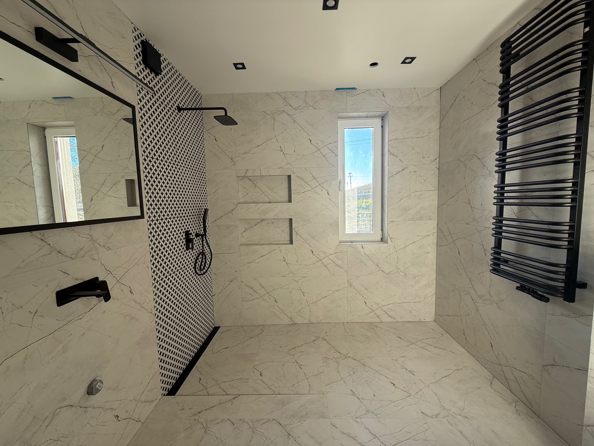 Wykończona łazienka z marmurowymi płytkami, czarną armaturą i grzejnikiem drabinkowym. Kabina prysznicowa z wzorzystą ścianą. Nowoczesny design z oknem.