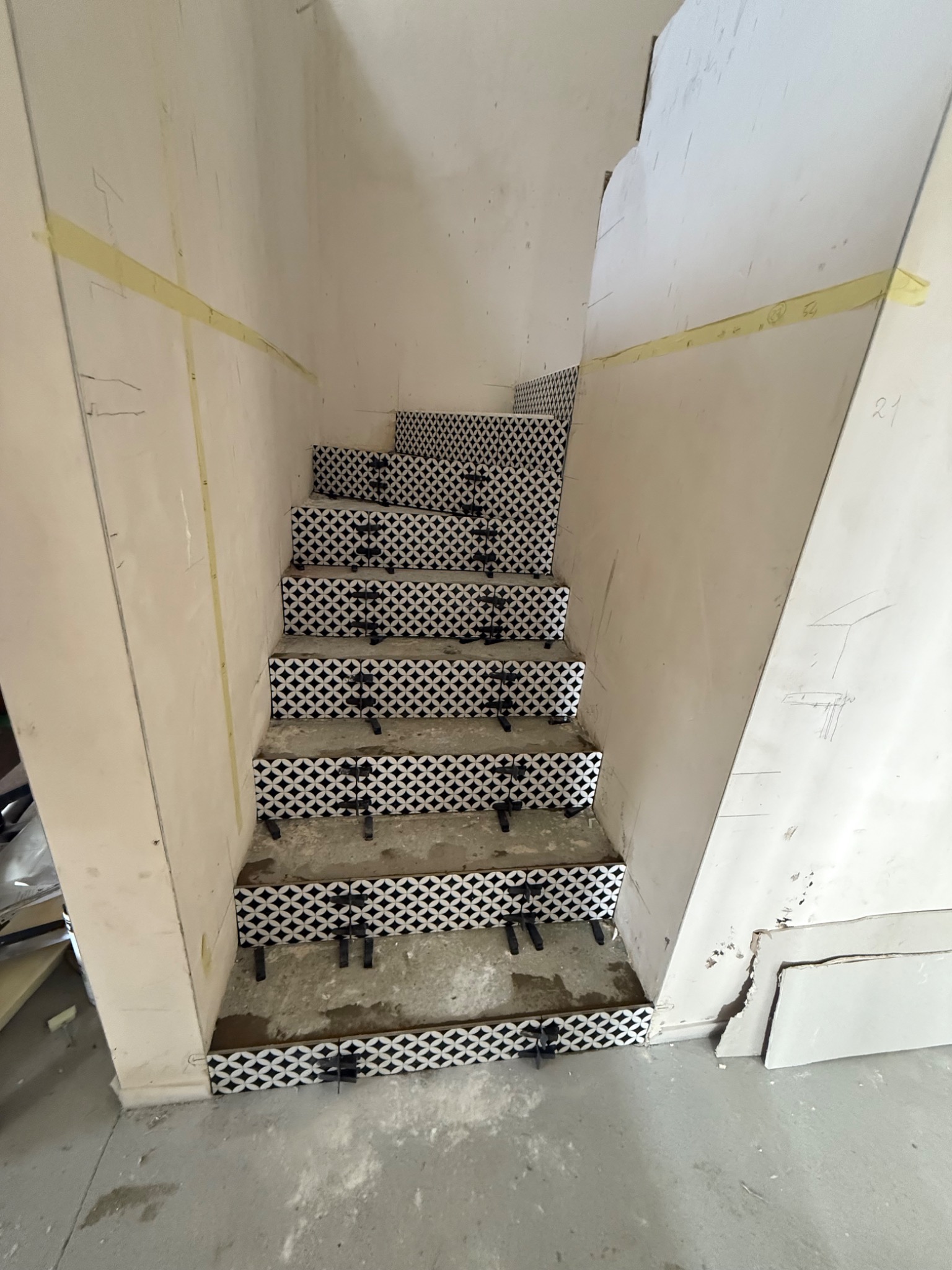 Schody w trakcie wykańczania z czarno-białymi płytkami, widoczne elementy mocujące. Ściany surowe, z taśmą malarską i oznaczeniami. Ujęcie z dołu schodów.
