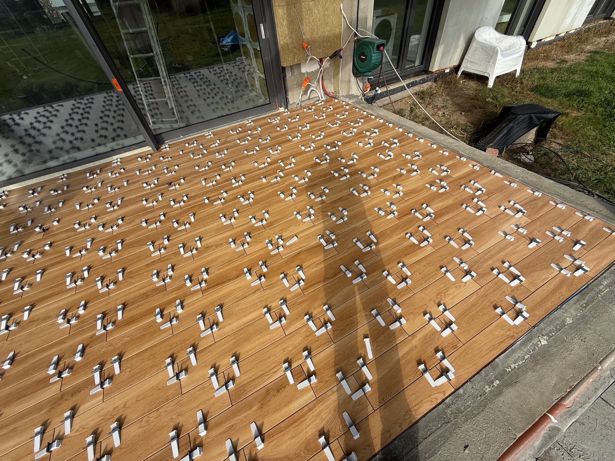 Układanie drewnopodobnych płytek na balkonie z użyciem systemu poziomowania. Widoczne białe klipsy i kliny. W tle okno tarasowe i elementy elewacji.