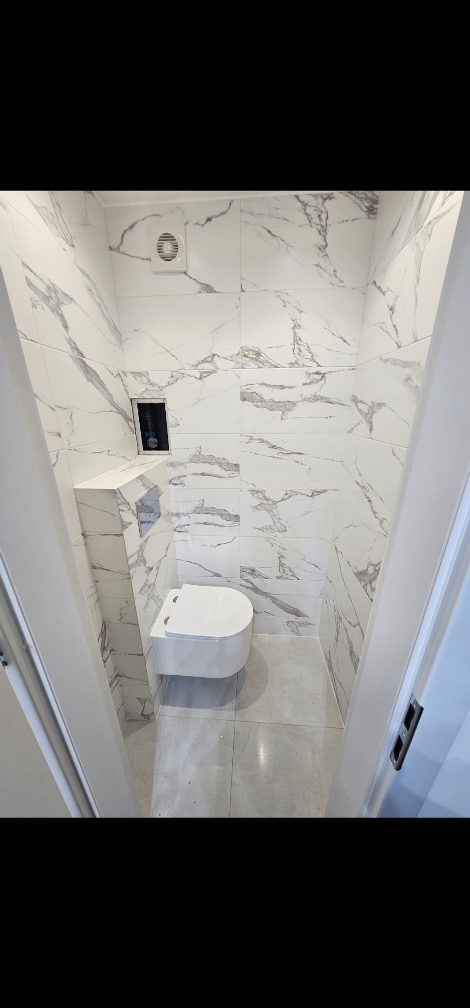Kompaktowa łazienka po remoncie: białe płytki imitujące marmur na ścianach i podłodze, podwieszana toaleta, wentylator. Widok zza futryny.