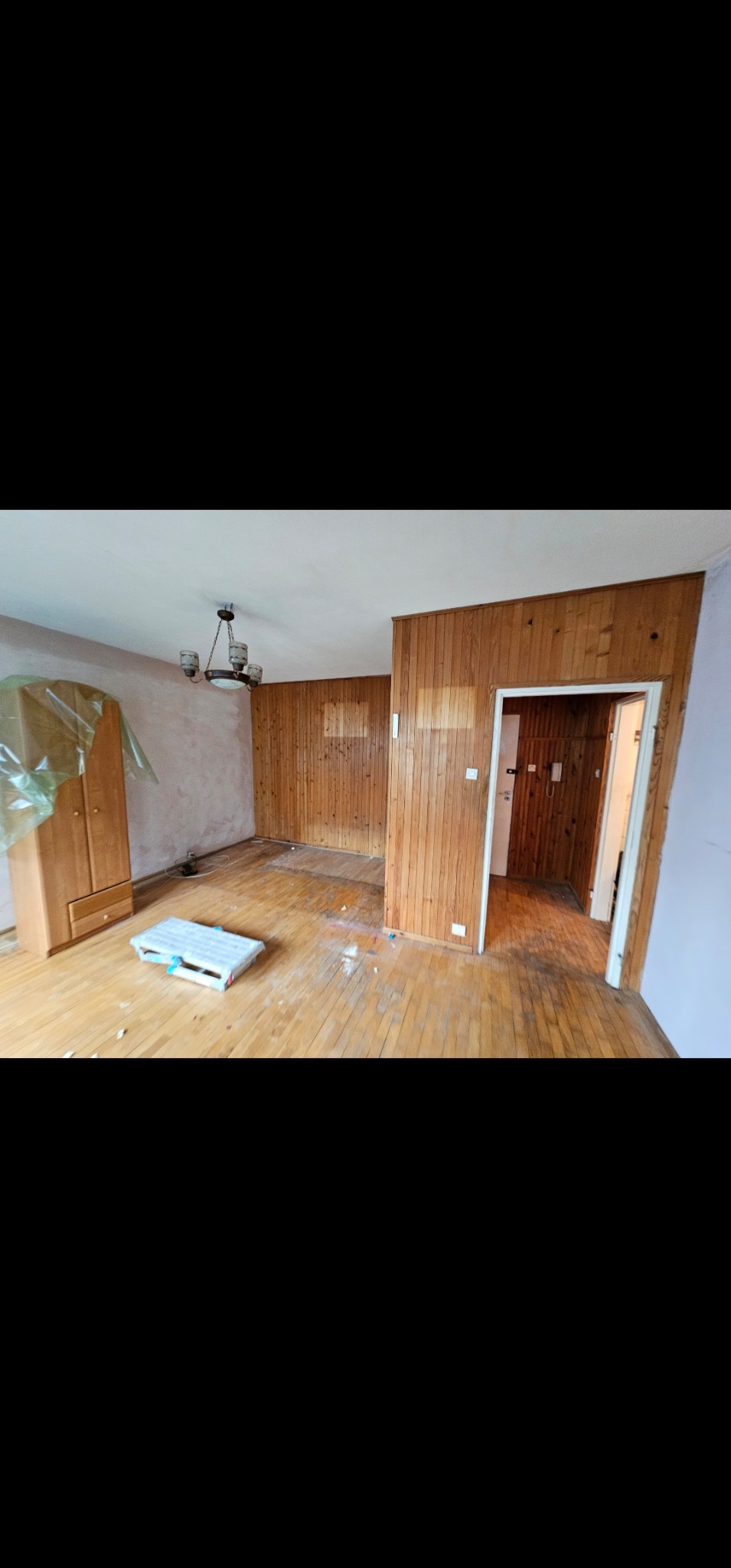 Pokój w trakcie remontu: drewniana boazeria na ścianach, częściowo zerwana, podłoga z desek, szafa zabezpieczona folią, widoczne ślady prac remontowych.