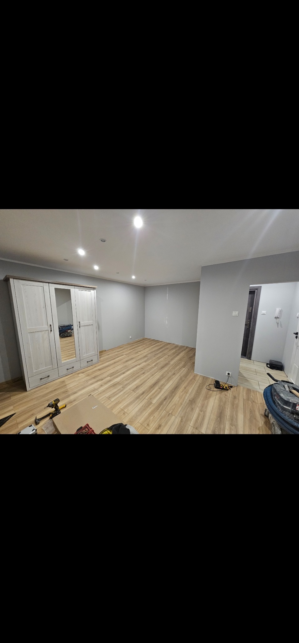 Jasne panele podłogowe ułożone w pokoju z szarą ścianą i białymi drzwiami. Widoczna szafa z lustrem i narzędzia pracy na podłodze. Oświetlenie sufitowe.