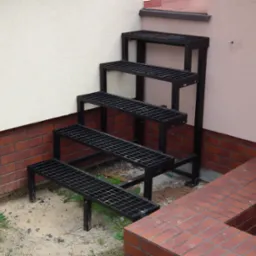 Zewnętrzne metalowe schody kratowe z czterema stopniami, umieszczone przy ścianie budynku z ceglaną podmurówką i piaskowym podłożem z zielenią.