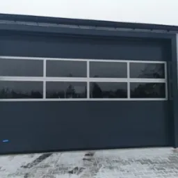 Szara brama garażowa segmentowa z poziomym oknem dzielonym na sześć części, zamontowana w hali przemysłowej, widoczny fragment ośnieżonego chodnika.