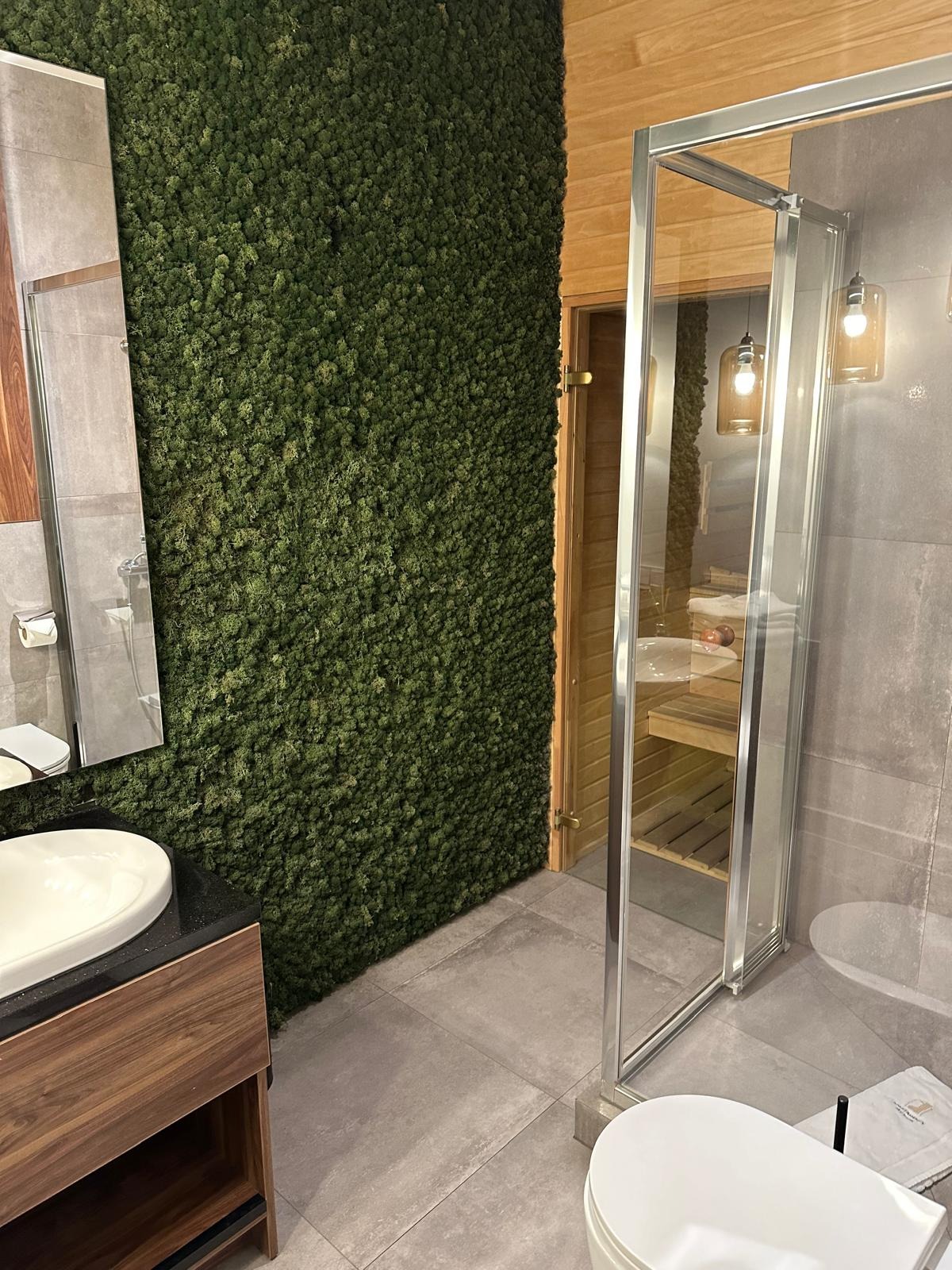 Nowoczesna łazienka z ścianą pokrytą mchem, kabiną prysznicową z widokiem na saunę, umywalką nablatową na szafce z drewnopodobnym fornirem i szarą płytką podłogową.