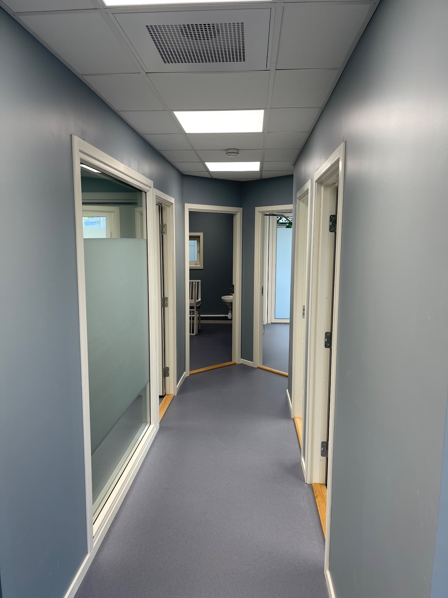 Korytarz biurowy z szarymi ścianami, białymi drzwiami i szarą podłogą, widok w głąb pomieszczeń z wyposażeniem.