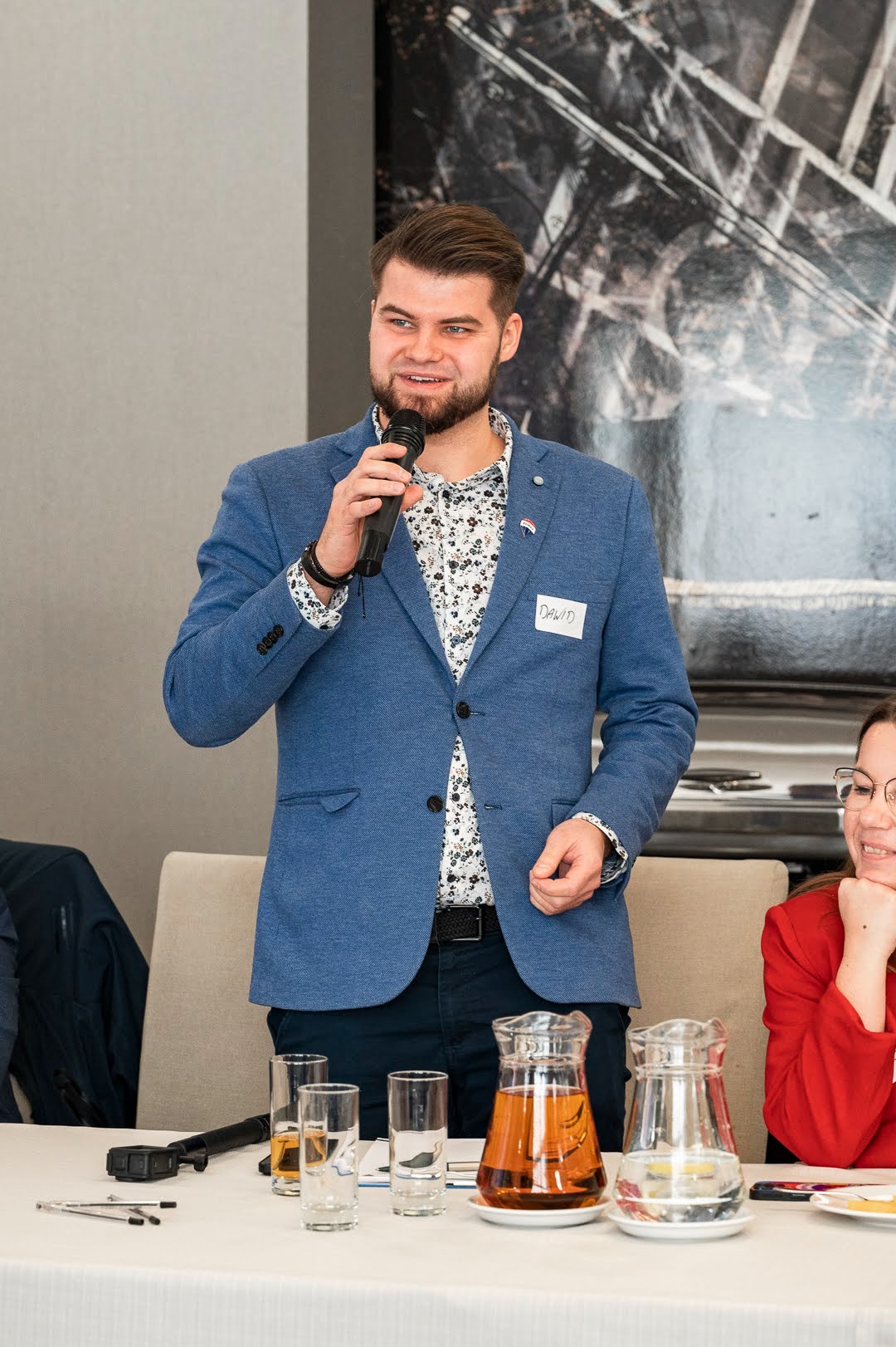 Mężczyzna w niebieskiej marynarce z identyfikatorem 'David' przemawia do mikrofonu na spotkaniu biznesowym, na stole szklanki z napojami i karafki.