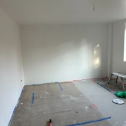 Przygotowane do malowania wnętrze pokoju z zabezpieczonymi taśmą podłogami i listwami, widoczny wałek malarski i drabina.