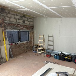 🏠 REMONTY & WYKOŃCZENIA WNĘTRZ 🏠 Jacek Sońta - Wnętrze w trakcie prac w Łodzi: widoczna surowa cegła, okno z PCV, częściowo zamontowana płyta na suficie i ścianach, drabiny, narzędzia i materiały budowlane.