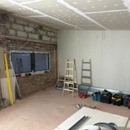 Wnętrze w trakcie prac w Łodzi: widoczna surowa cegła, okno z PCV, częściowo zamontowana płyta na suficie i ścianach, drabiny, narzędzia i materiały budowlane.