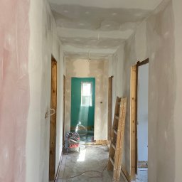 🏠 REMONTY & WYKOŃCZENIA WNĘTRZ 🏠 Jacek Sońta - Korytarz w trakcie prac wykończeniowych: ściany częściowo pokryte gładzią, widoczna drabina, szlifierka do gładzi i przewody na podłodze, w tle pomalowane na zielono pomieszczenie z oknem.