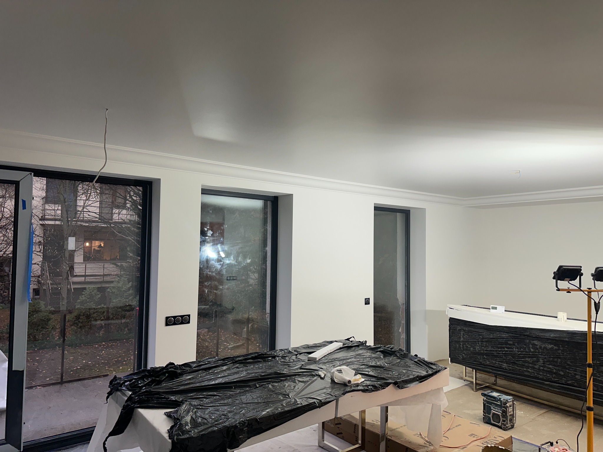 Wnętrze w trakcie prac wykończeniowych, zabezpieczone folią meble i podłoga, widoczne listwy przysufitowe, lampy budowlane i narzędzia.