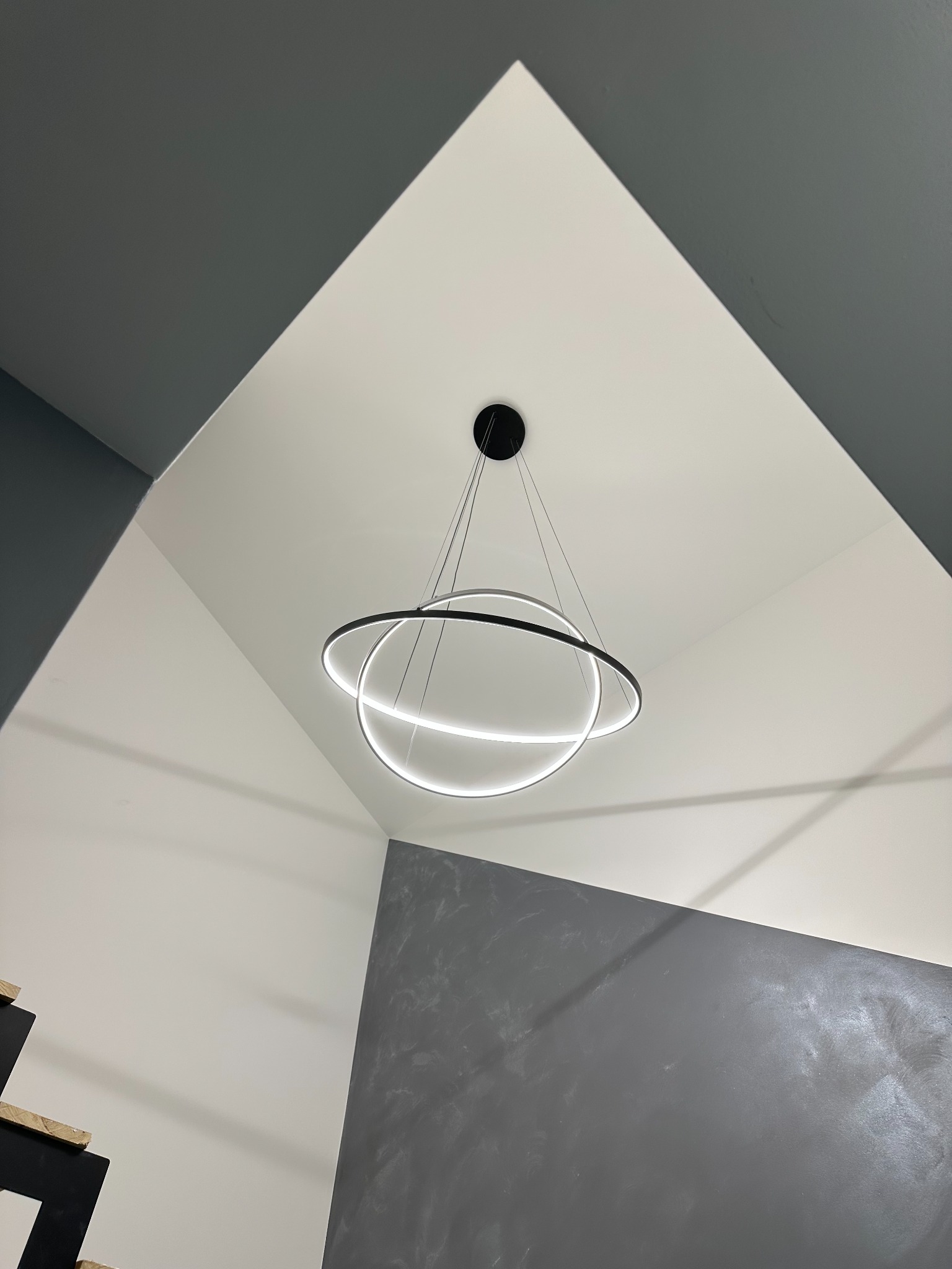 Nowoczesny, okrągły, ledowy żyrandol z dwoma pierścieniami zawieszony na cienkich linkach w rogu pokoju z białymi ścianami i szarą ścianą o fakturze betonu.