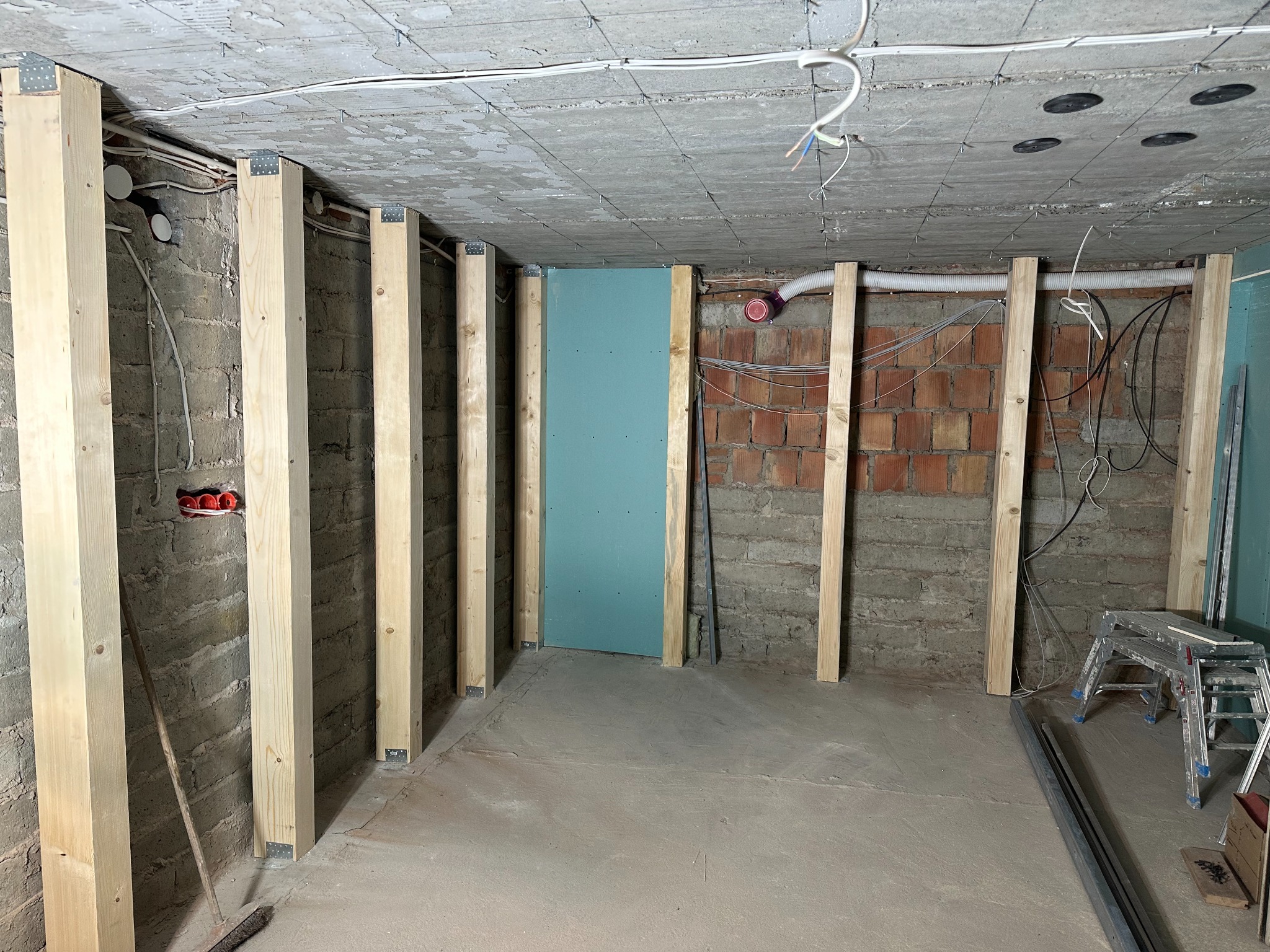 Surowa przestrzeń w trakcie prac budowlanych: drewniane słupki wzmacniające konstrukcję, widoczne instalacje elektryczne i wentylacyjne, fragment niebieskiej płyty oraz drabina w tle.