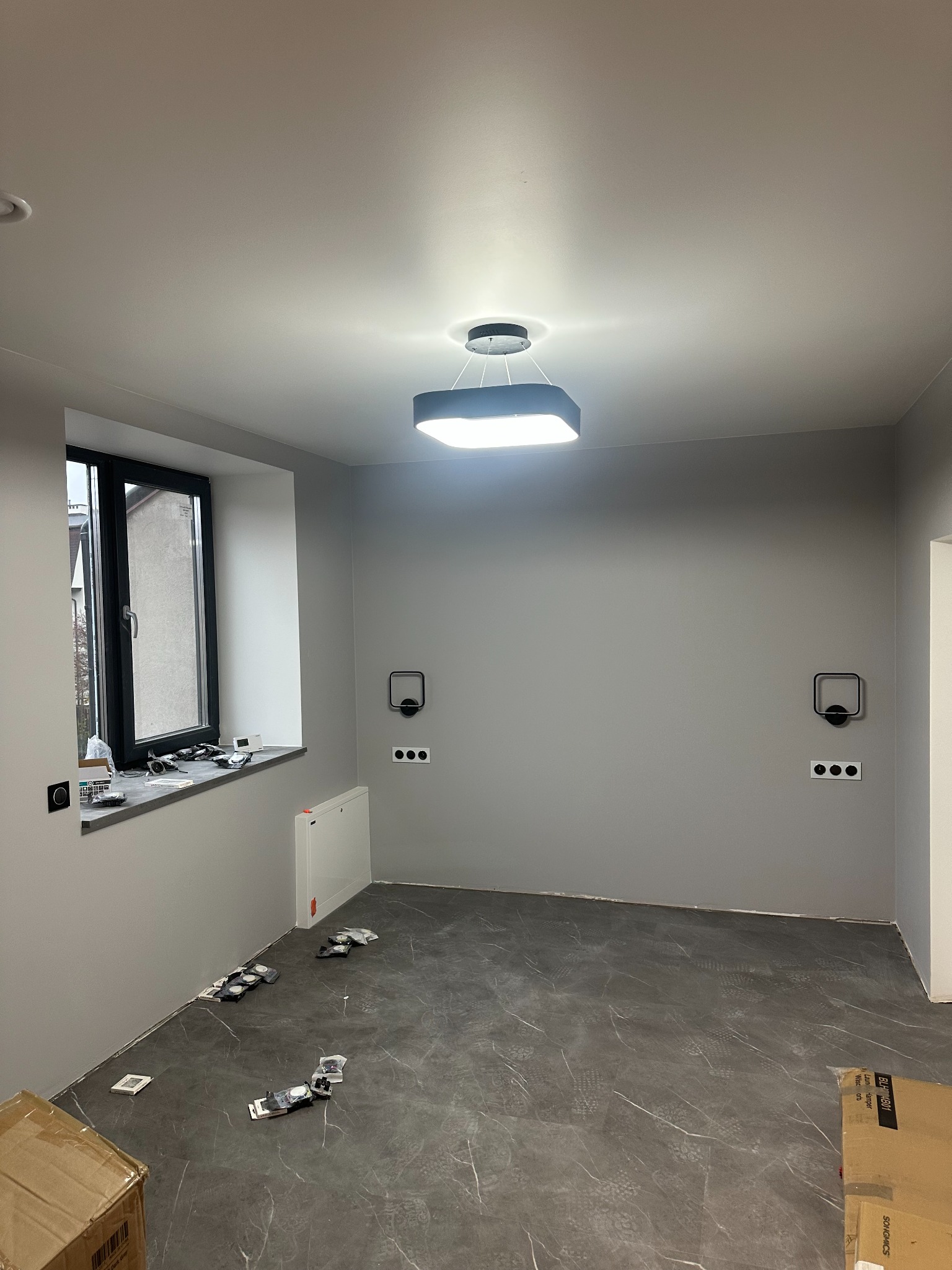 Szare wnętrze w trakcie wykańczania, z zamontowanym nowoczesnym oświetleniem sufitowym i ściennymi lampami, widoczne puszki po kleju na podłodze z imitacji marmuru.