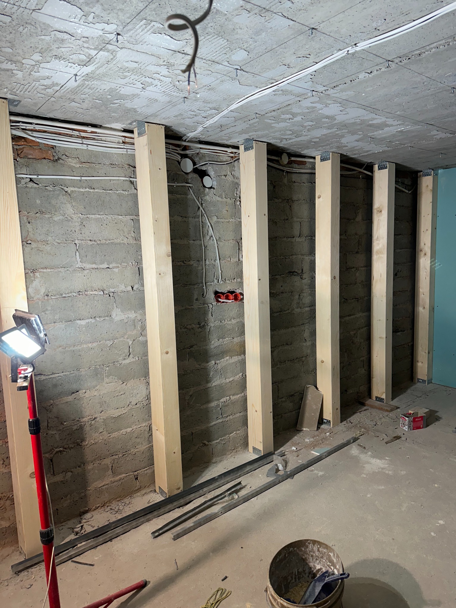 Surowa ściana z pustaków z zamontowanymi drewnianymi słupkami pod konstrukcję z płyt, widoczne przewody elektryczne i lampa robocza.