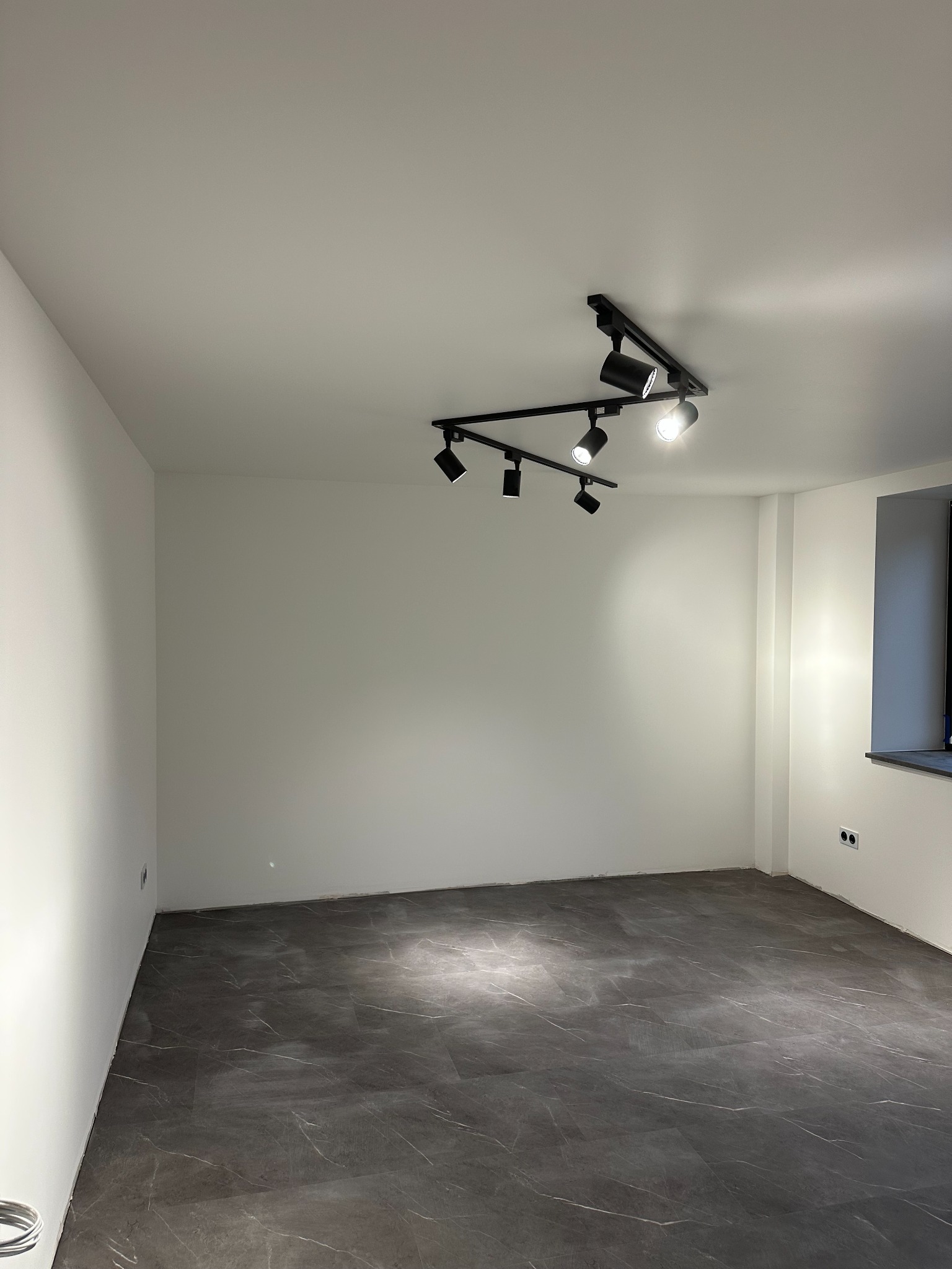 Pusty pokój po remoncie z ciemną podłogą imitującą marmur, białymi ścianami i nowoczesnym oświetleniem szynowym z reflektorami.
