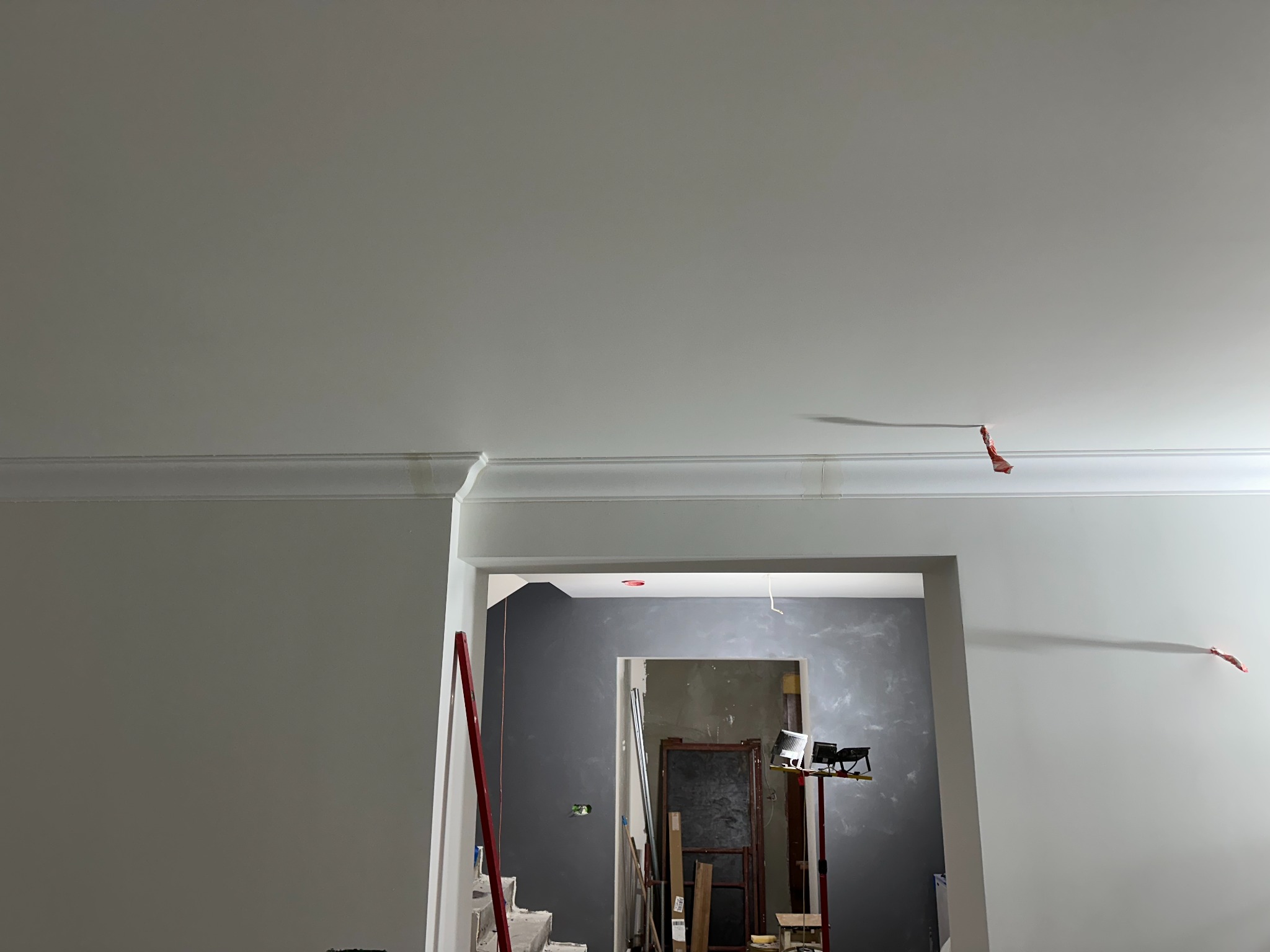 Wnętrze w trakcie malowania, widoczne ściany z dekoracyjną listwą przysufitową, szpachlowane ściany w innym pokoju widoczne w tle, lampa budowlana na statywie, poziomica oparta o ścianę.
