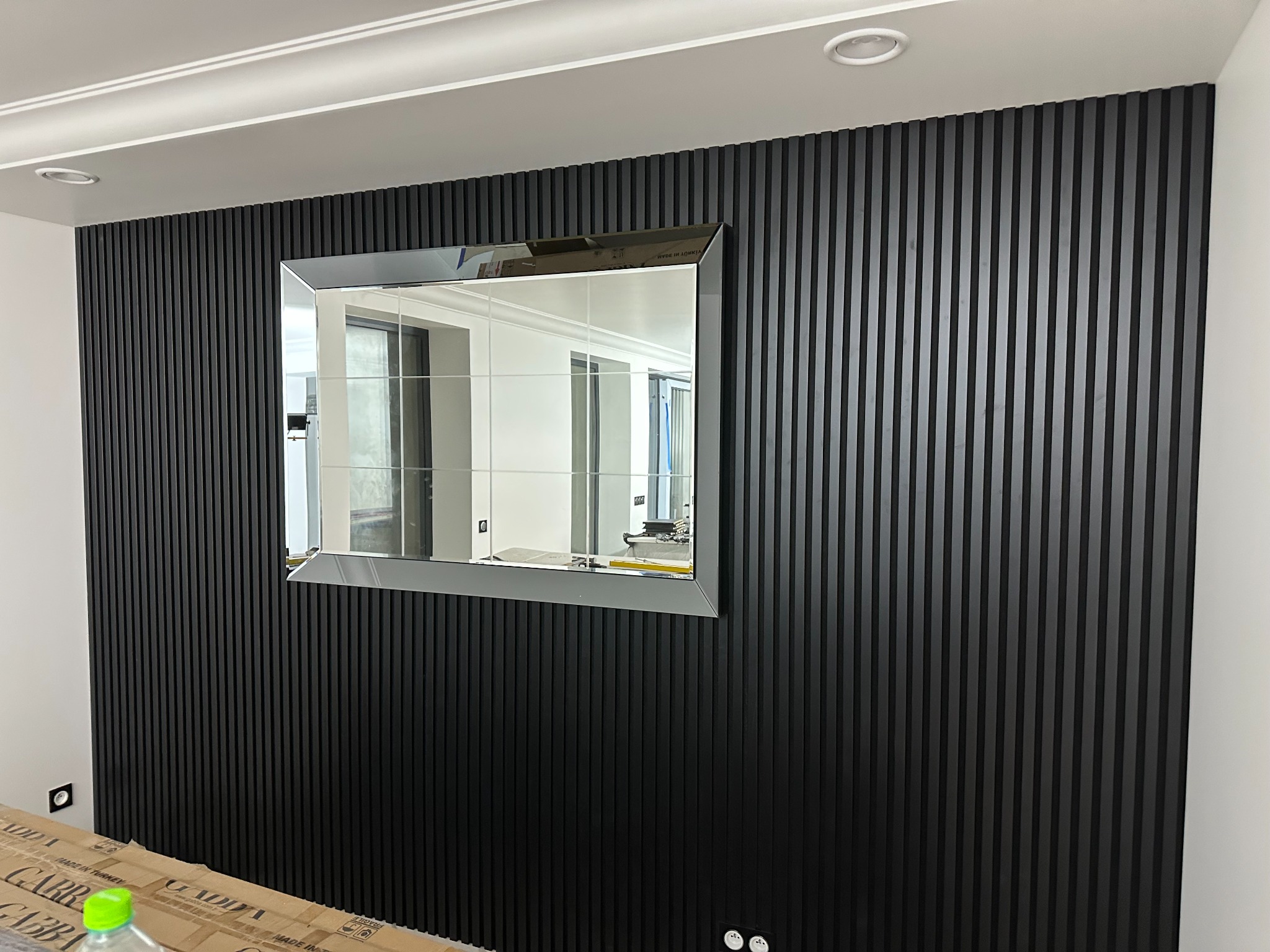 Ściana wykończona czarnymi, pionowymi lamelami z zamontowanym lustrem w srebrnej ramie, widoczne gniazdka elektryczne i fragment opakowania na podłodze.