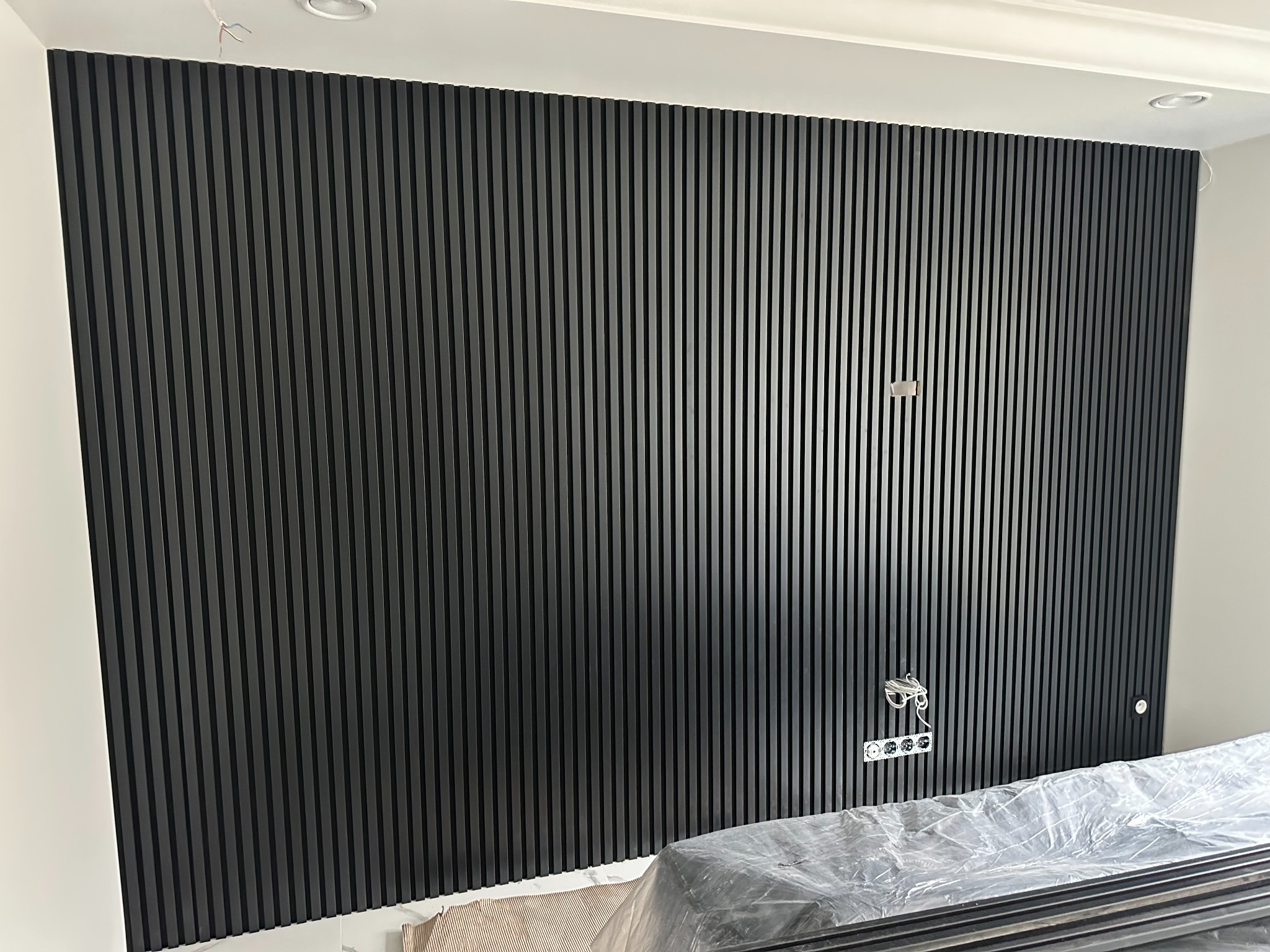 Ściana wykończona czarnymi panelami lamelowymi, gniazdka elektryczne na ścianie, elementy instalacji elektrycznej widoczne, panele zabezpieczone folią.
