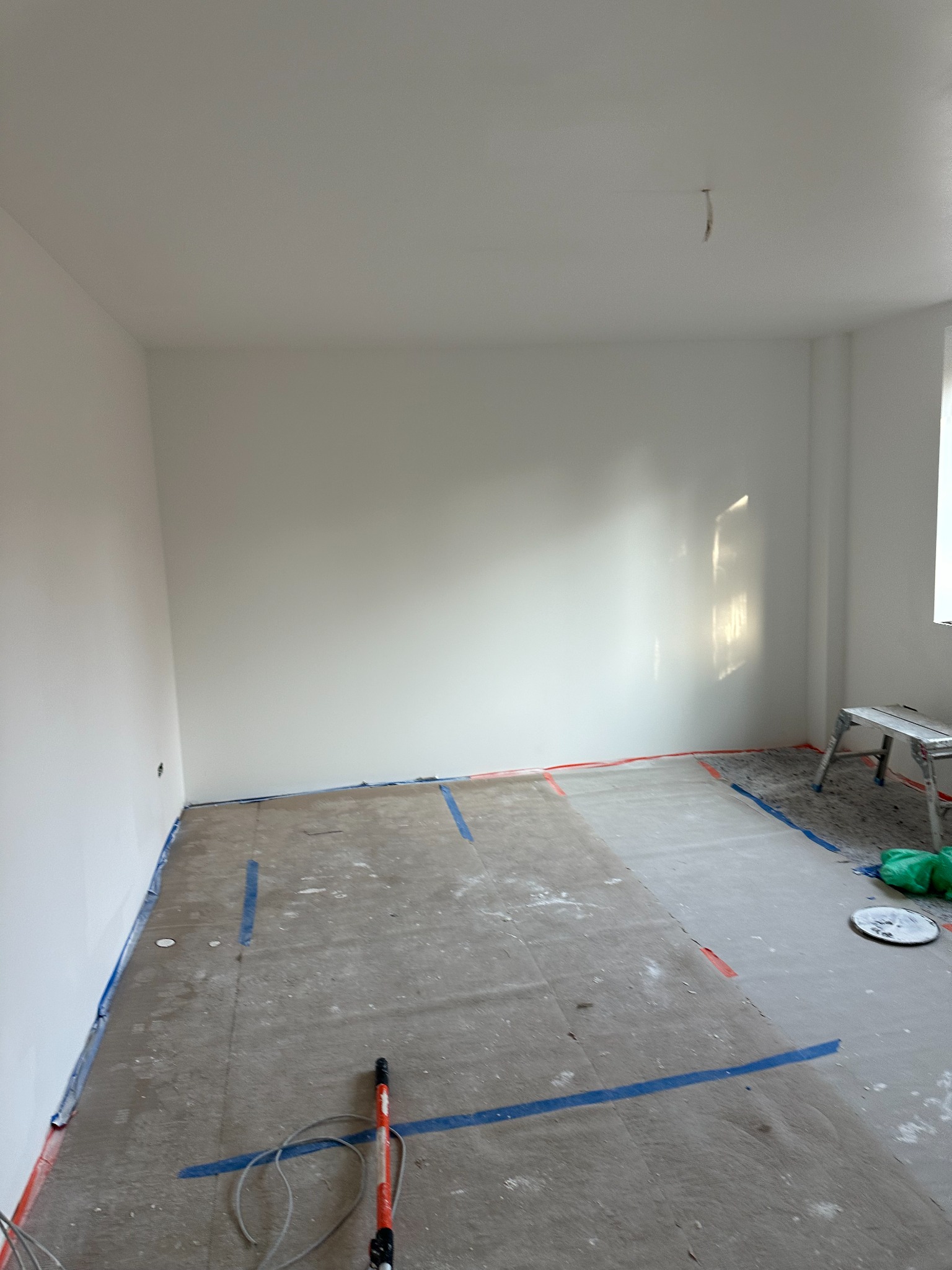 Przygotowane do malowania wnętrze pokoju z zabezpieczonymi taśmą podłogami i listwami, widoczny wałek malarski i drabina.