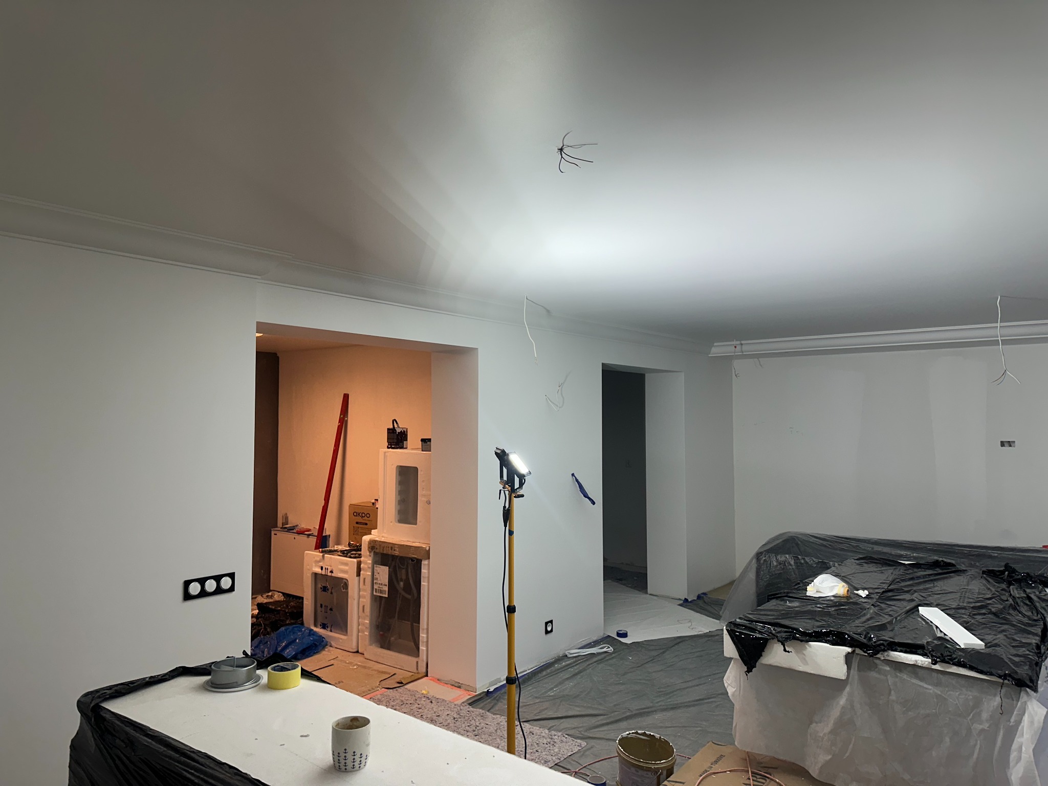 Pokój w trakcie remontu, ściany po szpachlowaniu i malowaniu na biało, widoczne zabezpieczenia folią, lampa budowlana i przygotowane materiały.