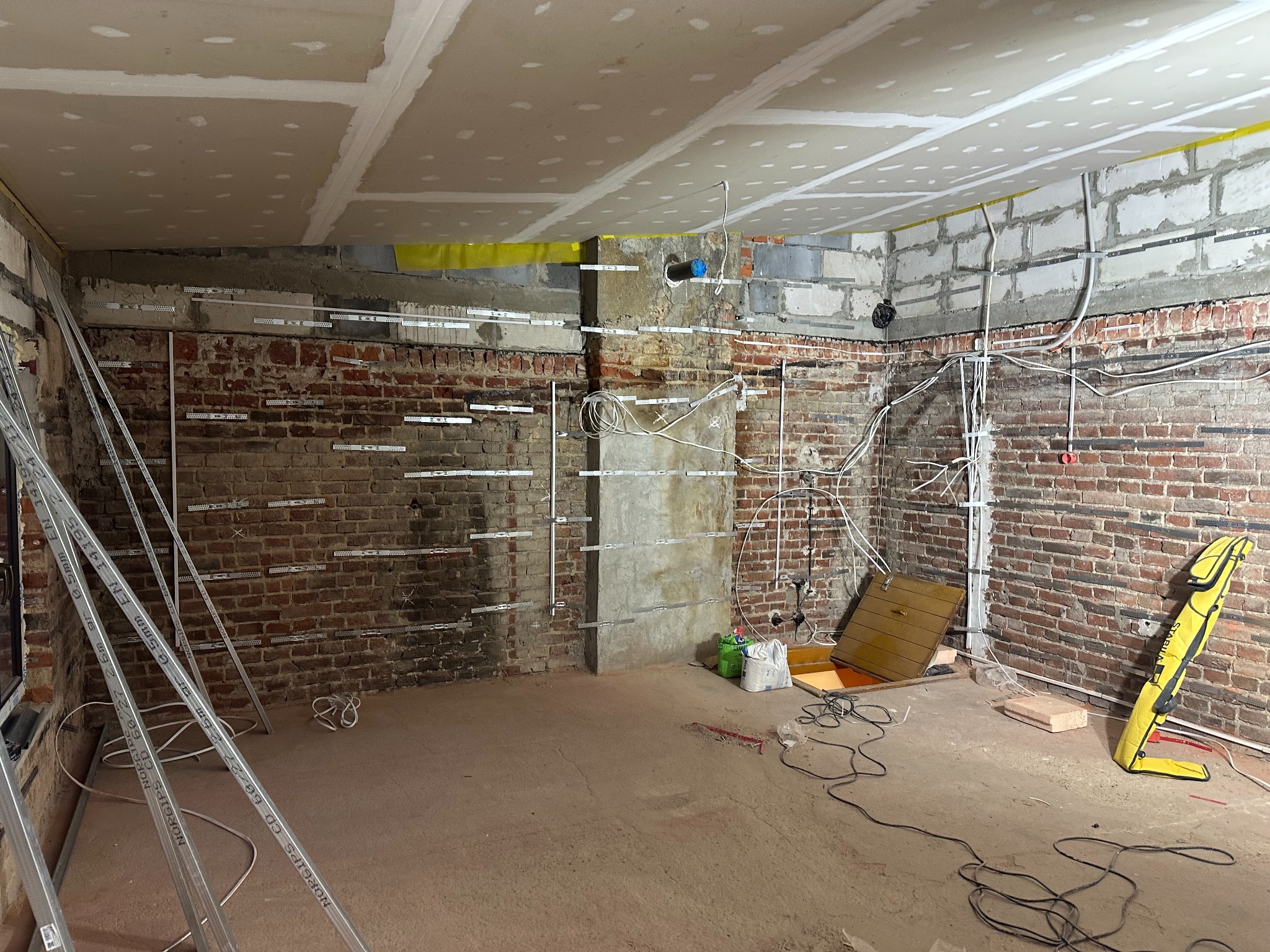 Wnętrze w trakcie prac remontowych: surowa cegła, instalacje elektryczne, częściowo zamontowane profile do płyt gipsowo-kartonowych na ścianach i suficie, widoczne przewody i narzędzia.
